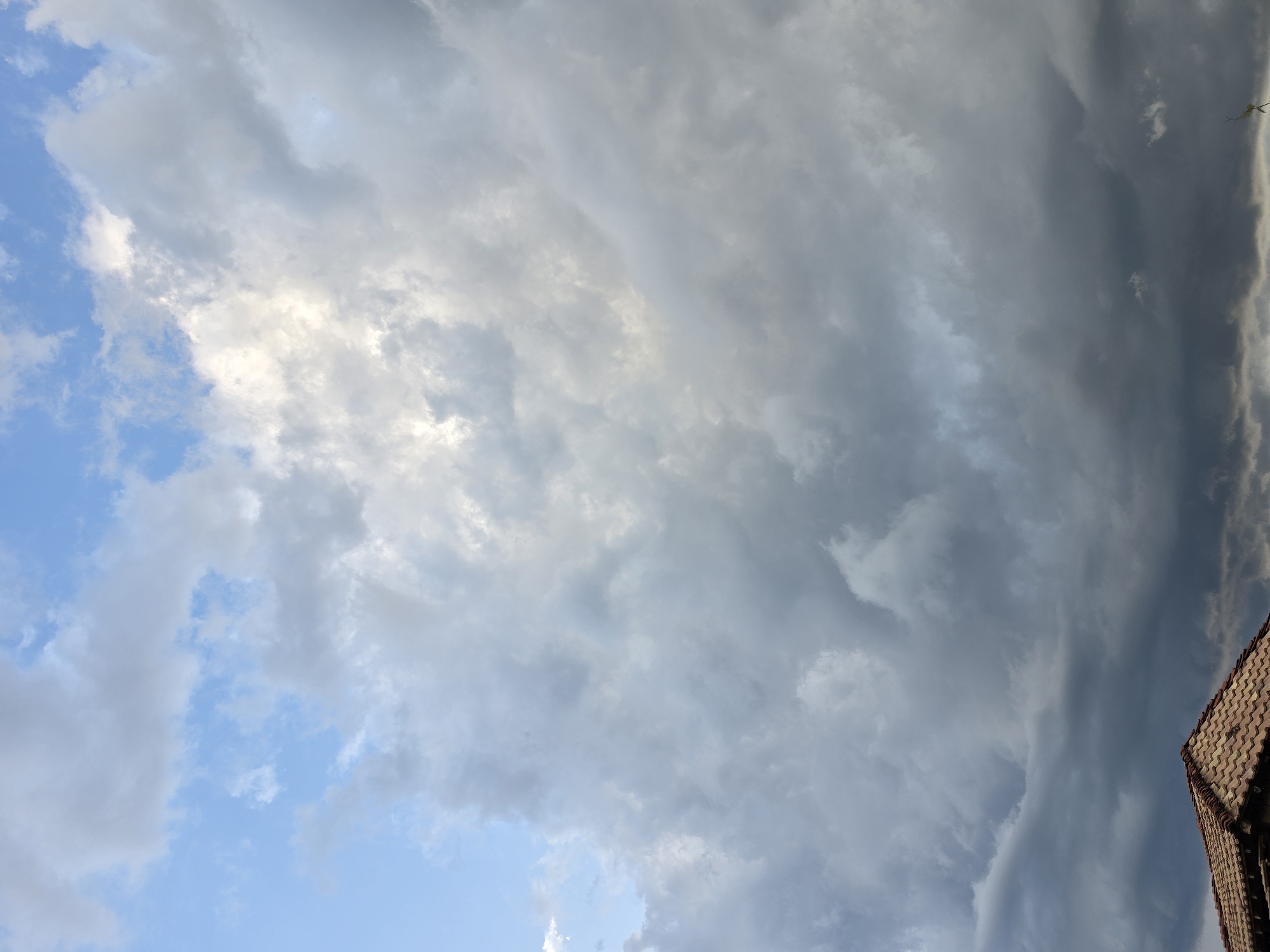 The image depicts a dramatic sky filled with a mix of light and dark clouds. The upper portion of the frame shows patches of blue sky interspersed with bright, fluffy clouds catching the sunlight. As the eye moves down, the clouds become denser and darker, suggesting an approaching storm or overcast conditions. In the lower left corner, the terracotta tiled roof of a building is visible, indicating an urban or suburban setting. The scene is devoid of any people or discernible action, presenting a natural, atmospheric view. The location is identified as Aleksandrovo, Bulgaria, suggesting this is a snapshot of the sky above that particular place.