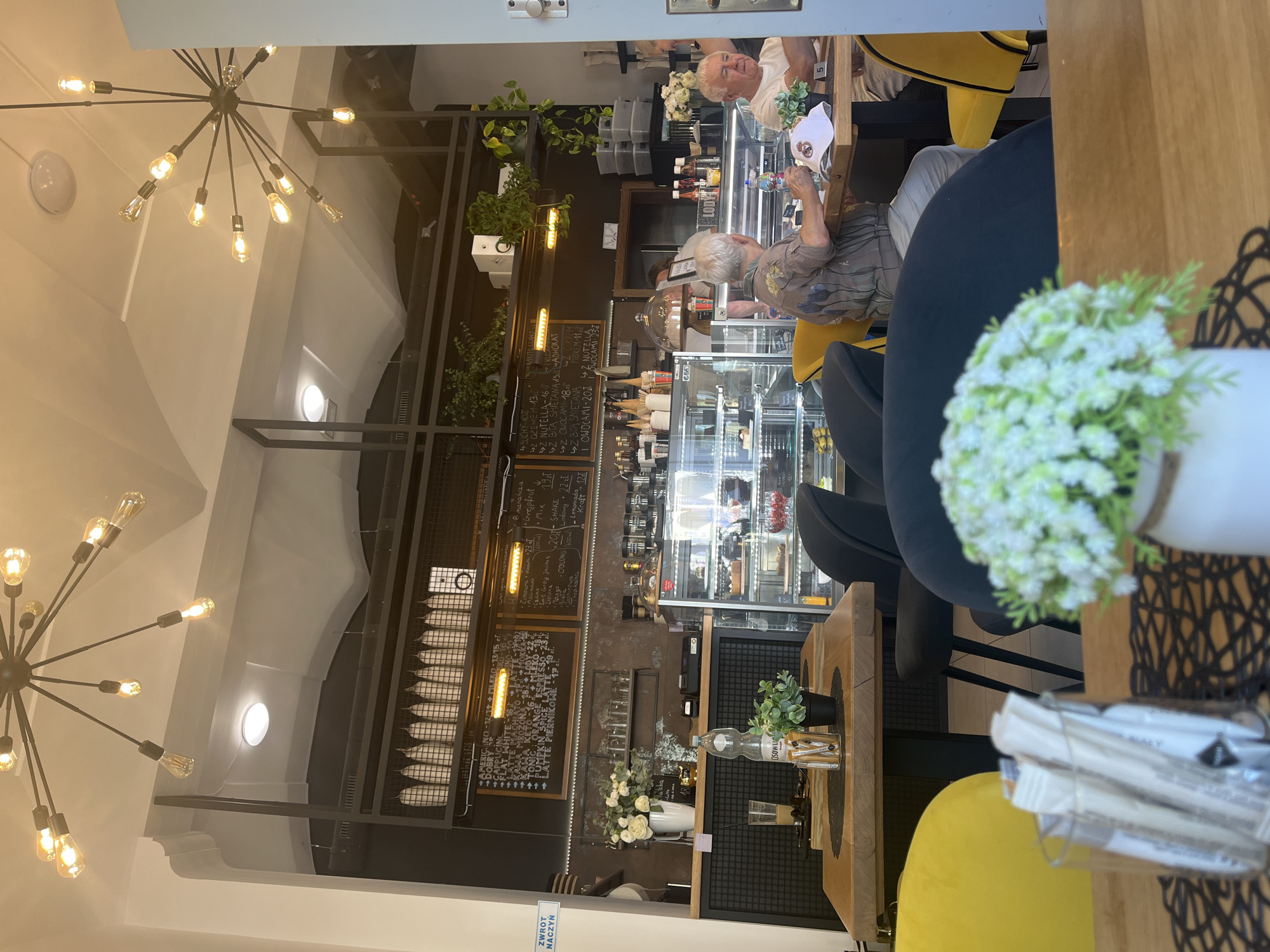 This image captures an indoor scene, likely a cafe or restaurant, with a focus on a display case filled with desserts and bottles. Two older individuals are seated at a table, one looking towards the display case and the other engaged with something on the table. The setting appears modern with industrial-style lighting fixtures and dark shelving units. There are plants and floral arrangements decorating the space. A prominent display case showcasing various cakes and pastries suggests a sweet treat establishment. The overall ambiance seems relaxed and casual.
Visible text on chalkboards includes menu items and prices, with Polish words like "NUTELLA," "SHAKE," and "OWOCAMI" (fruits) being discernible. The lighting suggests it could be daytime or evening, with warm artificial lights creating a cozy atmosphere. The presence of a bottle of "CISOWI" water provides a specific brand cue. The scene conveys a sense of everyday activity in a dining establishment.