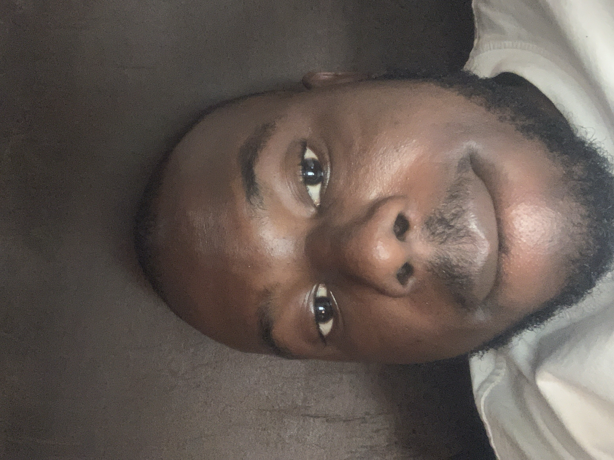 The image depicts a close-up shot of a man's face. He has dark skin, dark eyes, and a short beard. The background is out of focus and shows a dark surface, possibly a couch or a wall, and a glimpse of a white shirt. The image provides no information related to the city of Jos, Nigeria.