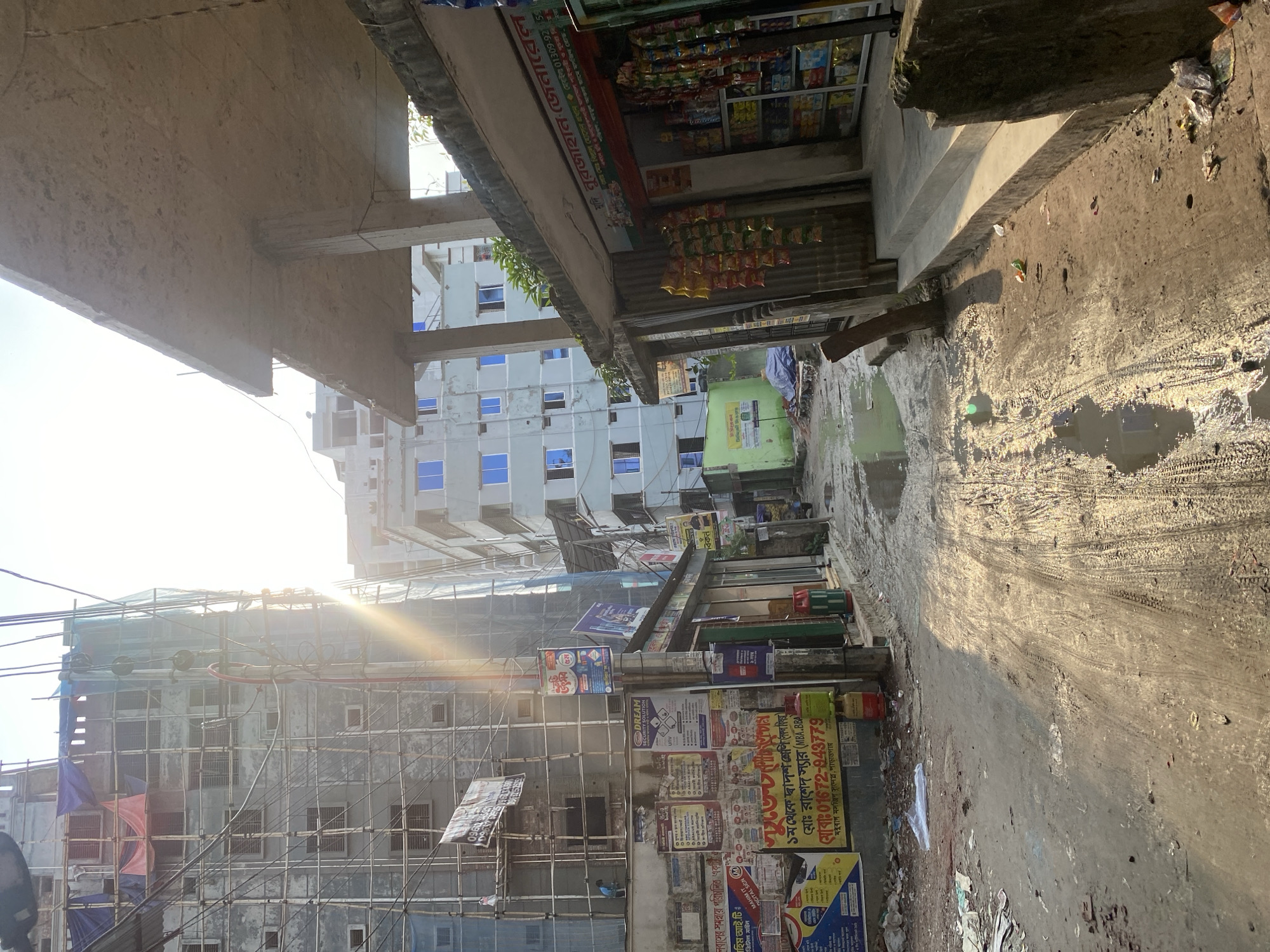 The image depicts a narrow, somewhat unclean street in Dhaka, Bangladesh. On one side, a multi-story building is under construction, shrouded in scaffolding and partially covered in tarps. Numerous posters and advertisements are plastered on the lower sections of buildings along the street. Small shops line the street, selling various goods, including snacks and stationery. The street itself is a mix of wet patches and dry areas, marked with tire tracks. Sunlight filters through the buildings, creating a bright contrast.