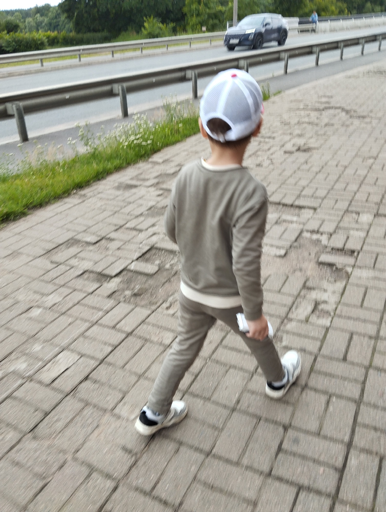 A child walks on a brick walkway next to a road with a guardrail. The child is wearing a grey sweatshirt, grey pants, a white mesh baseball cap with a red accent, and white sneakers with black accents. The child is holding a white object in their right hand. In the background, a dark SUV is driving on the road, and trees are visible beyond the road. The walkway has cracked and uneven paving stones. The image appears to be taken in Kingisepp, Russia, based on the provided context.
