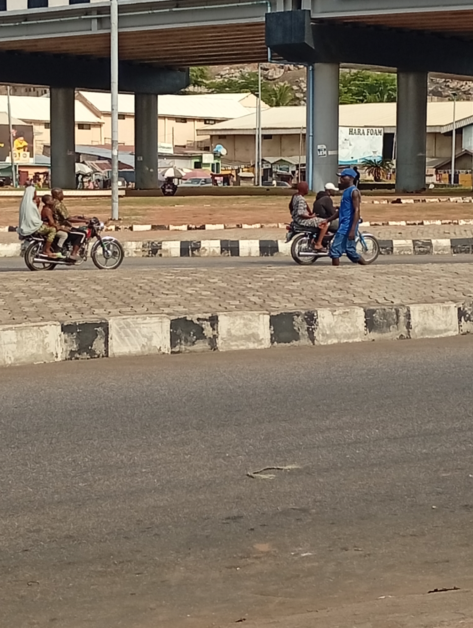 The image, taken in Lokoja, Nigeria, depicts a street scene under an overpass. Two motorcycles are visible, each carrying passengers. The first motorcycle has a driver and two passengers, one with a white head covering. The second motorcycle has a driver and one passenger, with a person walking beside it holding a bicycle. In the background are buildings, one with "HARA FOAM" displayed on its side. The foreground consists of an asphalt road and a concrete curb with black and white painted stripes. The weather appears sunny and the environment is urban.