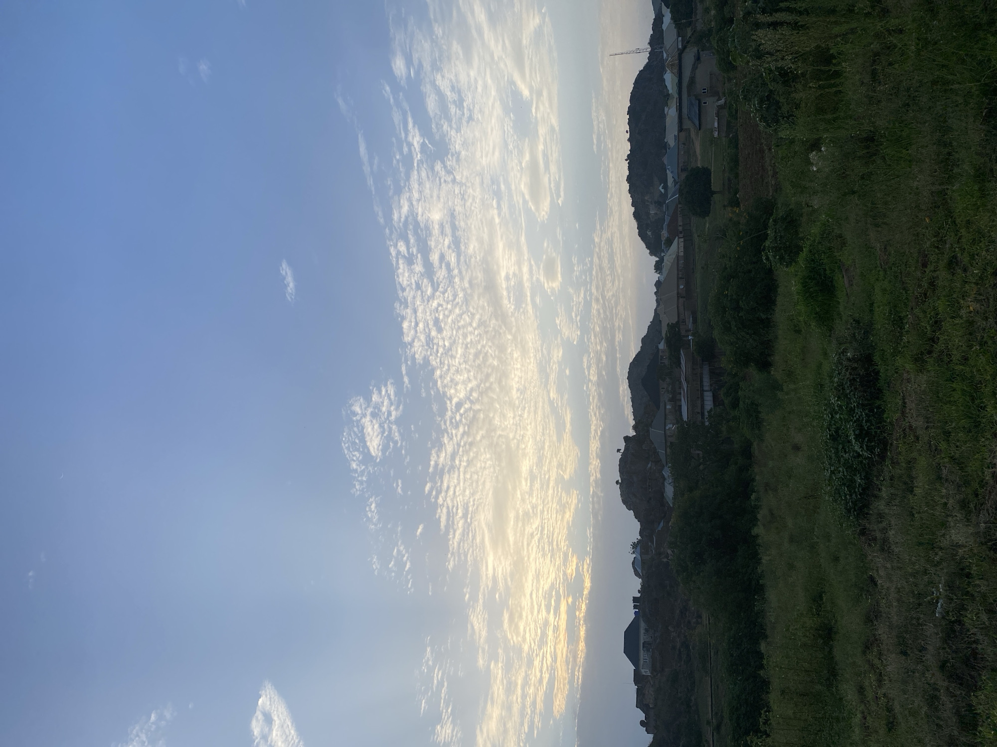 The image captures a landscape in Gura Top, Nigeria, at either sunrise or sunset. The sky dominates the view, displaying a combination of clear blue and dense, patterned altocumulus clouds that stretch across the majority of the frame. The clouds are illuminated with a soft, golden light.
The terrain on the right side of the image consists of a hillside covered in vegetation, with a mix of grasses and shrubbery. Several houses are nestled along the slope, blending into the natural environment. In the distance, a communications tower is visible amidst the buildings on the hilltop. The overall impression is one of a rural area with a scattered settlement.