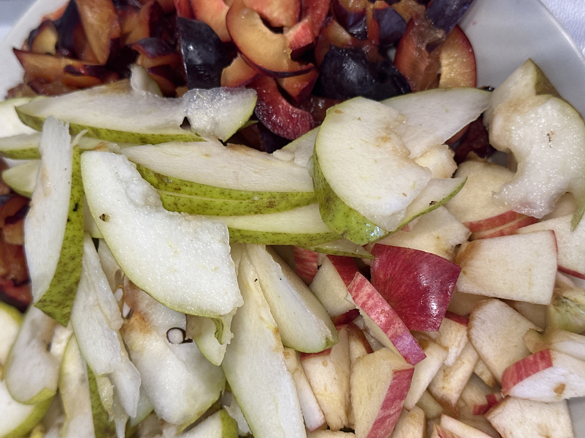 A close-up, overhead shot shows a plate of sliced fruit. The fruit is divided into three distinct sections. On the left are chopped apples, with their red skin visible in thin strips against the white flesh. In the center are sliced pears, their green skin contrasting with the pale flesh. To the right are sliced plums, with deep purple and reddish-brown hues. The fruit appears to be prepared for a meal or dessert. There is no discernible location in Nevinnomyssk, Russia, or any other environmental context visible in the image.