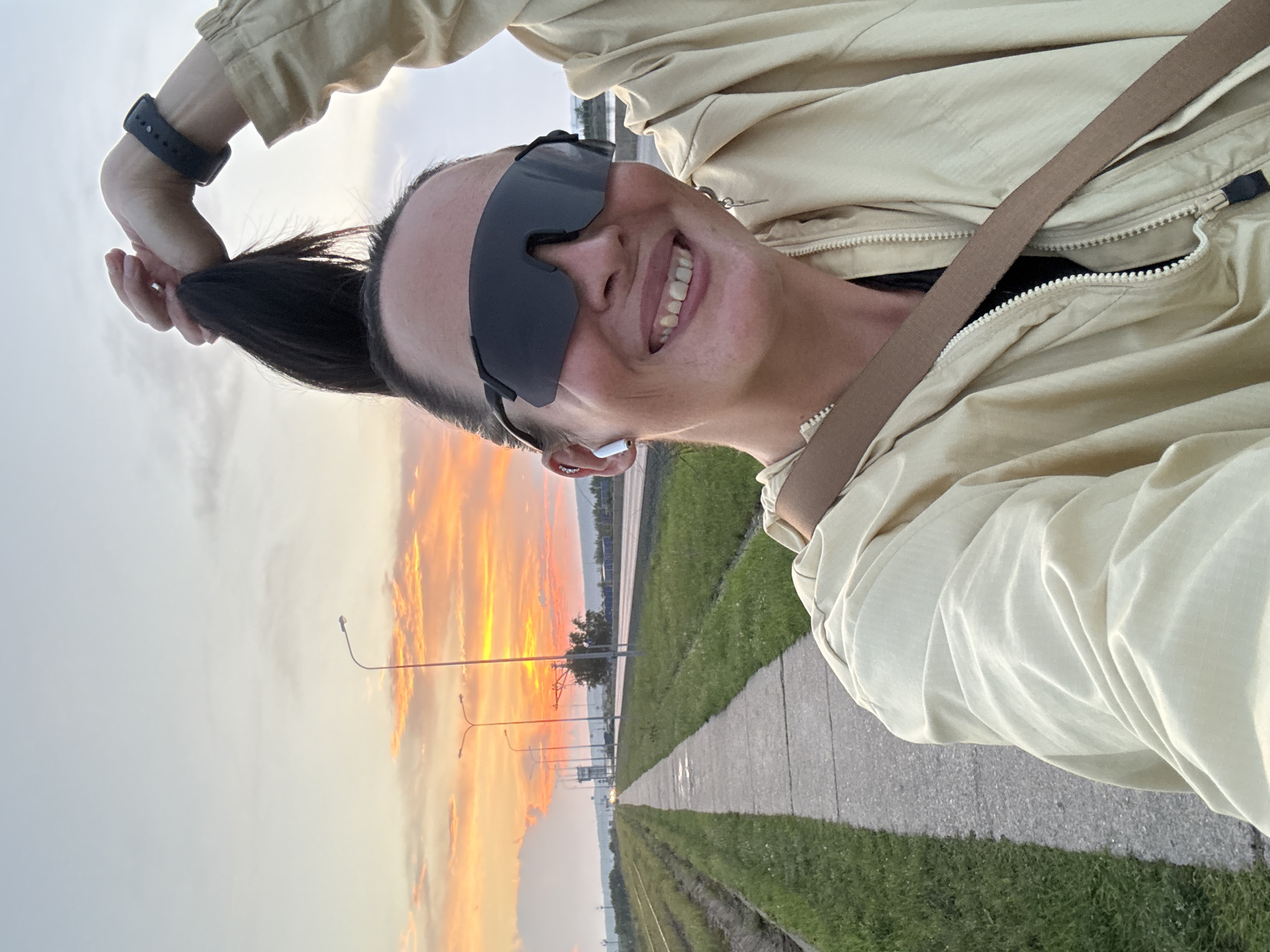 A woman with dark hair pulled into a high ponytail, wearing large black sunglasses, a light yellow hooded jacket, and a smartwatch on her left wrist, is taking a selfie outdoors during sunset. She is smiling and holding her ponytail with her right hand. She also has a beige strap visible across her jacket. The background shows a sidewalk next to a grassy area, a road with streetlights, and a dramatic orange and yellow sky with clouds. The setting appears to be an urban or suburban area with infrastructure like roads and railway tracks visible. This scene is consistent with locations along the Kama River in Chaikovsky, Russia, where such expansive sunset views over industrial or transportation corridors are common.