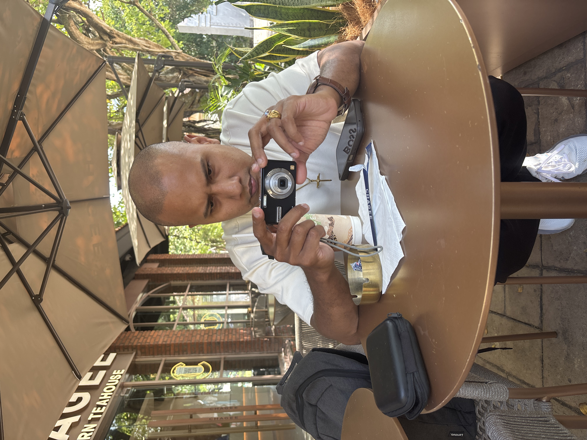In Kuta Selatan, Indonesia, a bald, medium-skinned man sits at a round, brown table under a beige umbrella outside a teahouse. He wears a light-colored t-shirt, black pants, and white sneakers. He holds a black digital camera to take a photo of himself. On the table are a gold ashtray with cigarette butts, a disposable cup, a black case, a smartphone, and a napkin. A gray backpack rests on a chair to his left, and plants are visible in the background. The teahouse facade features brickwork and large windows displaying the interior.