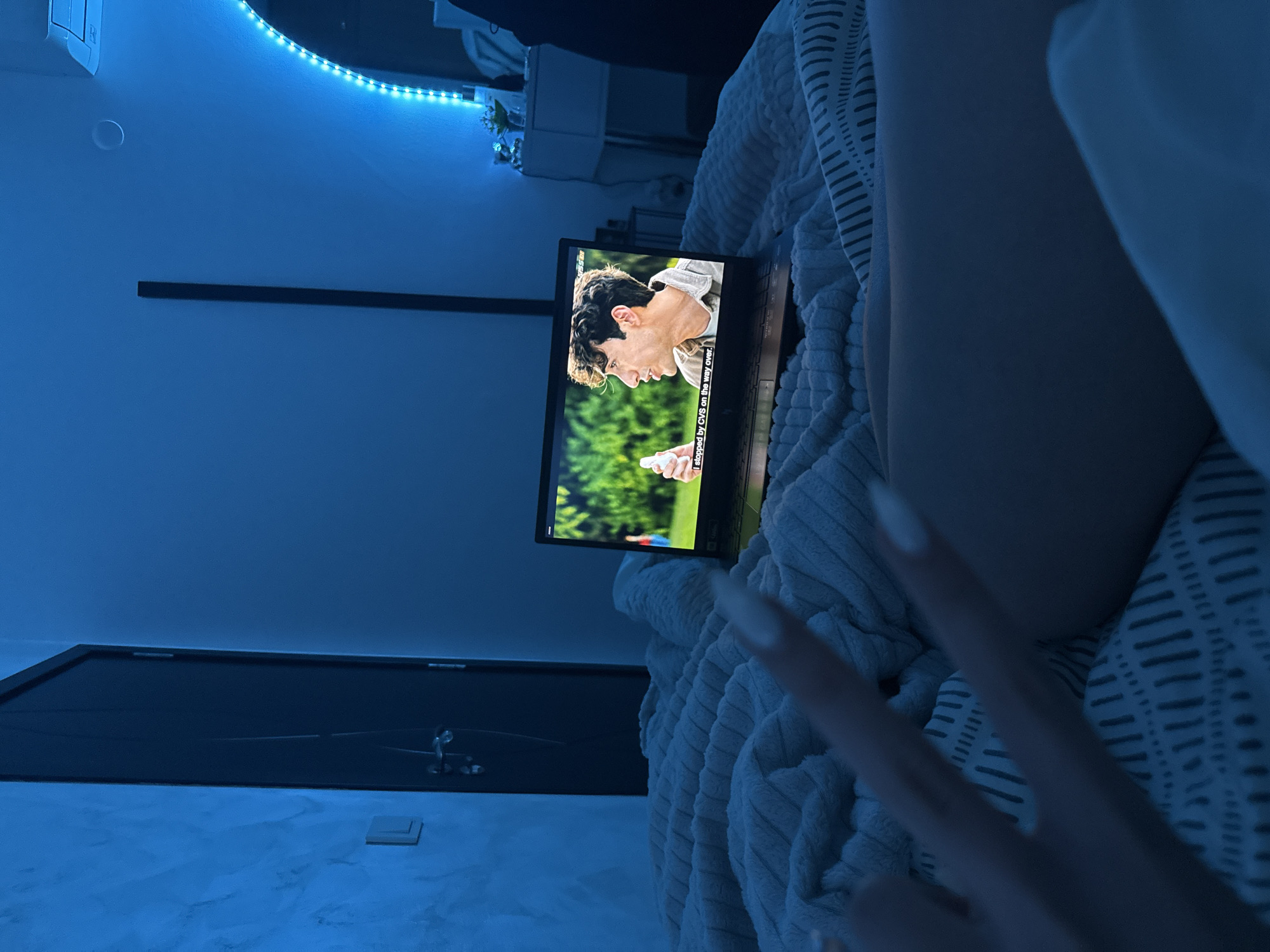 A person is in bed watching a laptop. The laptop screen shows a man and subtitles: "I stopped by CVS on the way over". The room has LED strip lights, a shelf with decorative flowers, and a dark door. A hand with long nails is raised in the foreground. The scene is illuminated in blue light. The location cannot be identified as Blagoevgrad, Bulgaria.