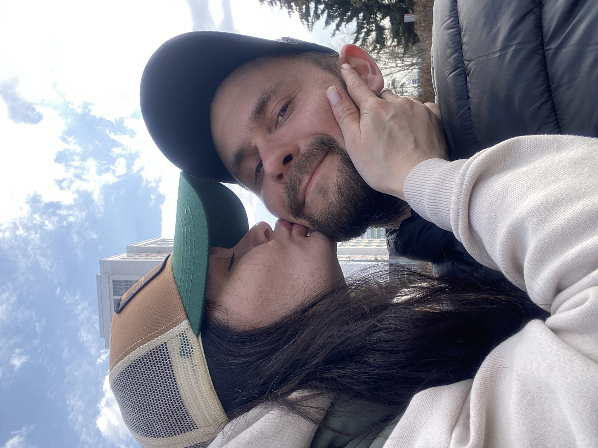 Two people are close together, possibly kissing. The person on the right has dark hair, a beard, and is wearing a black baseball cap. Their right hand is up near their face, touching the other person's face. The person on the left has long dark hair and is wearing a tan and green baseball cap. They are wearing a light-colored sweater. In the background, there is a building visible against a sky with clouds. It is not possible to identify the location as Kazan, Russia from the image provided.