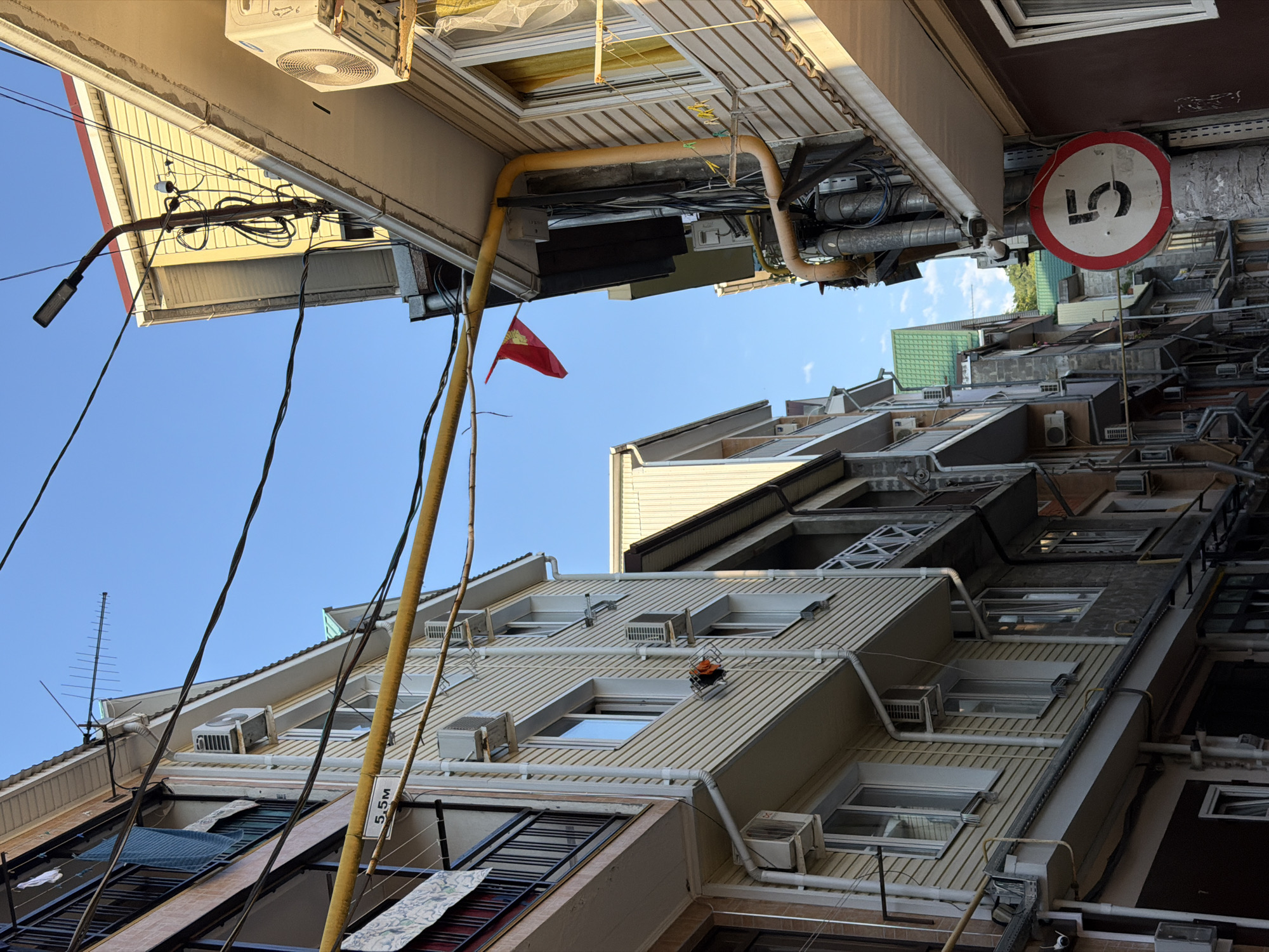 The image depicts a view looking upwards at the facades of buildings in a dense urban setting, likely a residential area. The perspective is skewed, creating a sense of looking up from a narrow street or alley. The buildings are multi-story, with many windows and air conditioning units visible. There are numerous electrical wires and pipes running along the exteriors of the buildings, creating a somewhat cluttered appearance.
A red flag with a yellow emblem, possibly a national or organizational flag, is seen flying from one of the buildings. A circular traffic sign with a red border and a white center featuring the number "5" is also visible on one of the buildings, suggesting a speed limit or some other numerical designation. Laundry is hung out to dry on some of the buildings, indicating domestic activity. The sky is clear and blue, suggesting a sunny day. The overall impression is of a vibrant, lived-in urban environment with many signs of human habitation and infrastructure. There is also a sign with "5.5m" written on it, possibly indicating a measurement. The time of day appears to be late afternoon or early evening, given the angle of the light.