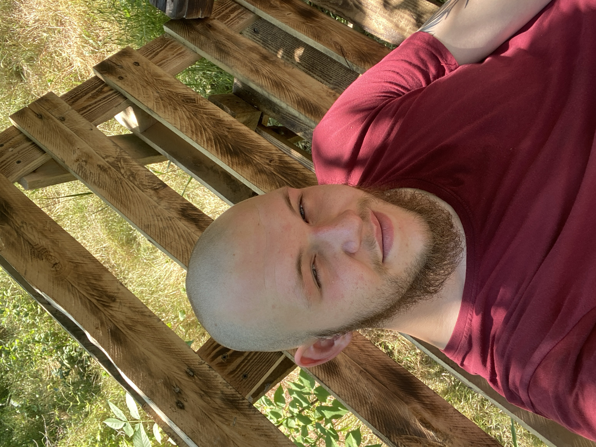 In Makiivka, Ukraine, a young man with a shaved head and a beard is pictured outdoors, seated on a wooden structure. He is wearing a maroon t-shirt and is looking towards the camera with a slight smile. The background consists of green foliage and grass, with the sunlight casting shadows on the wooden planks behind him. The wooden structure appears to be a bench or part of a larger seating arrangement.
