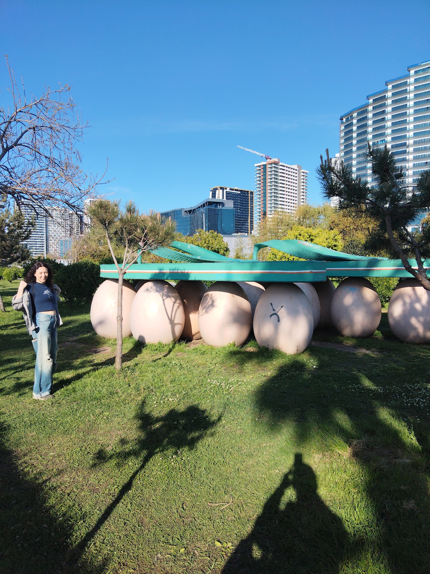 The image shows a woman standing in a grassy area in Batumi, Georgia. In the background is a large, whimsical sculpture composed of large, light-colored egg shapes topped by a green, oversized sandal. The backdrop includes modern high-rise buildings, including one with a crane indicating ongoing construction. The sky is clear and blue. A young tree stands in front of the sculpture. The shadow of the person taking the picture is visible in the foreground.