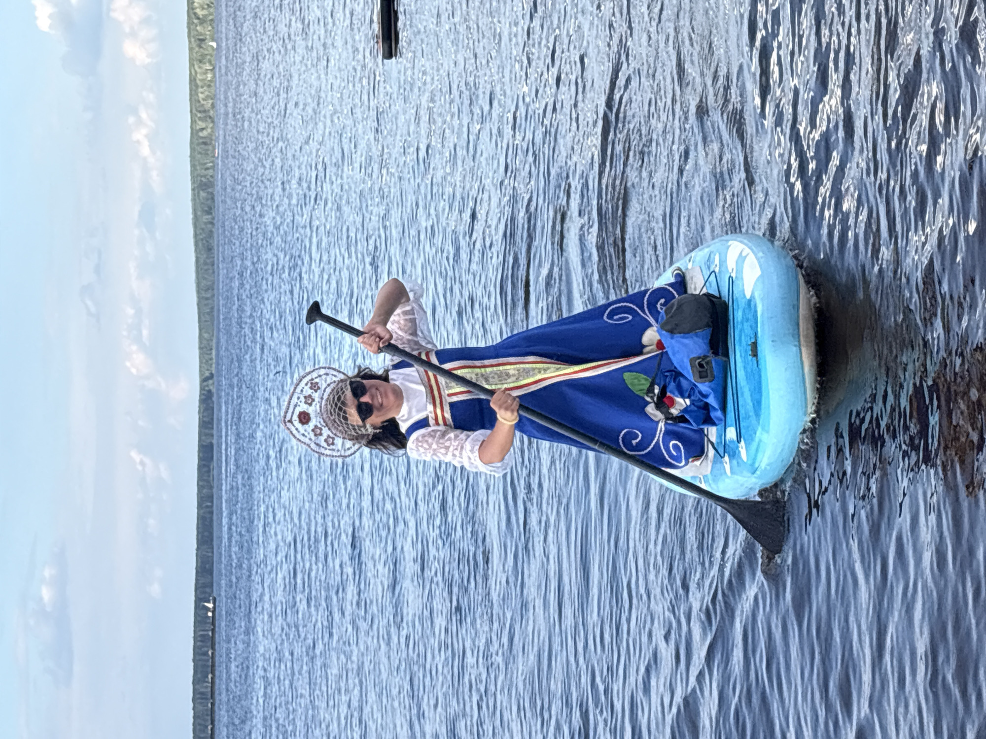 A woman is paddleboarding on a body of water, dressed in a traditional Russian costume with a kokoshnik headpiece and a blue sarafan. She is holding a paddle and smiling. The water is calm with small ripples. In the background, there is a tree-lined shore and a cloudy sky with some boats on the water. The scene appears to be taking place on the Kama River, near the city of Chaikovsky, Russia, as indicated by the geographical context of traditional Russian attire and the presence of a wide river consistent with the Kama.