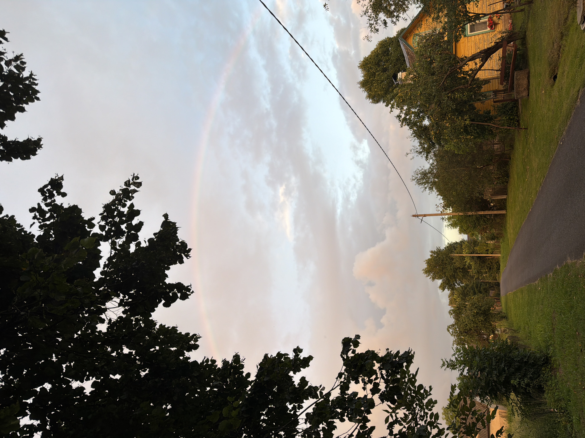 A dimly lit shot captures a rainbow arching across a sky with scattered clouds, illuminated by the soft glow of twilight. Silhouetted against the sky, a large tree with dark leaves occupies the left side of the frame. A paved road winds through a rural landscape, bordered by green grass and trees. On the right side of the road, a vibrant yellow house stands with its windows reflecting the fading light. Power lines stretch across the sky, adding a touch of urban infrastructure to the natural scene. The overall atmosphere is serene and tranquil, suggesting a peaceful evening in a quiet countryside setting.