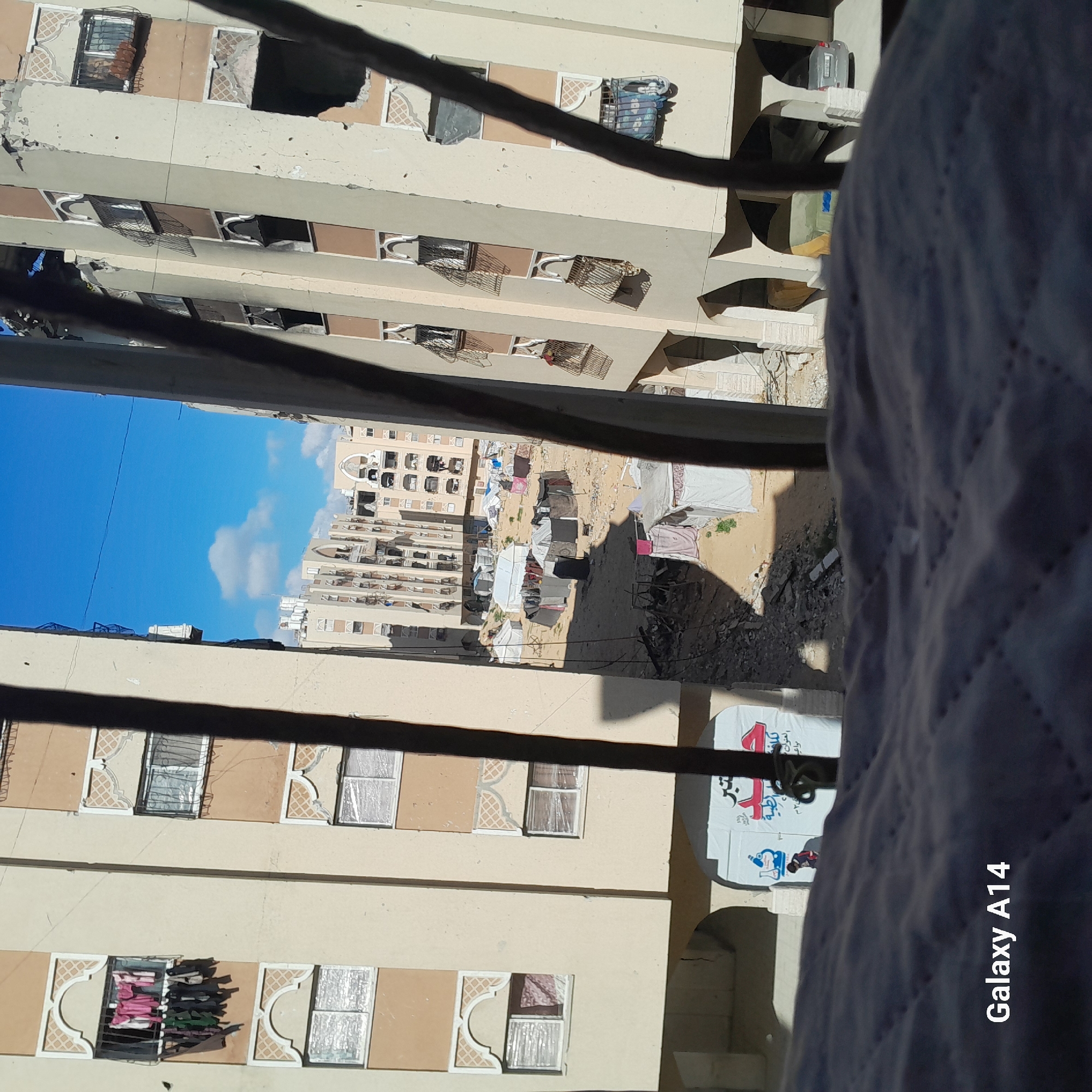 Here is a detailed description of the image:
The image appears to be taken from a high vantage point, possibly a balcony, overlooking a scene of damage and displacement in an urban area. The structures in the foreground and middle ground are multi-story buildings, many with visible damage to windows, walls, and balconies. Some windows are boarded up or covered with makeshift materials.
In the open area between the buildings, there are numerous temporary shelters constructed from cloth, plastic sheeting, and other available materials. These shelters suggest a displaced population living in basic conditions. The ground appears to be bare earth or sand.
The sky is visible through a gap between the buildings, showing a clear blue sky with some clouds. This creates a strong contrast between the devastation on the ground and the peacefulness of the sky.
The vantage point is partially obscured by what appears to be a railing or fence and a large, blurred object in the foreground, possibly part of the structure where the photo was taken. There's also a sign visible on one of the buildings.
Based on the scene's general characteristics, it suggests the aftermath of conflict or a natural disaster in an urban environment.