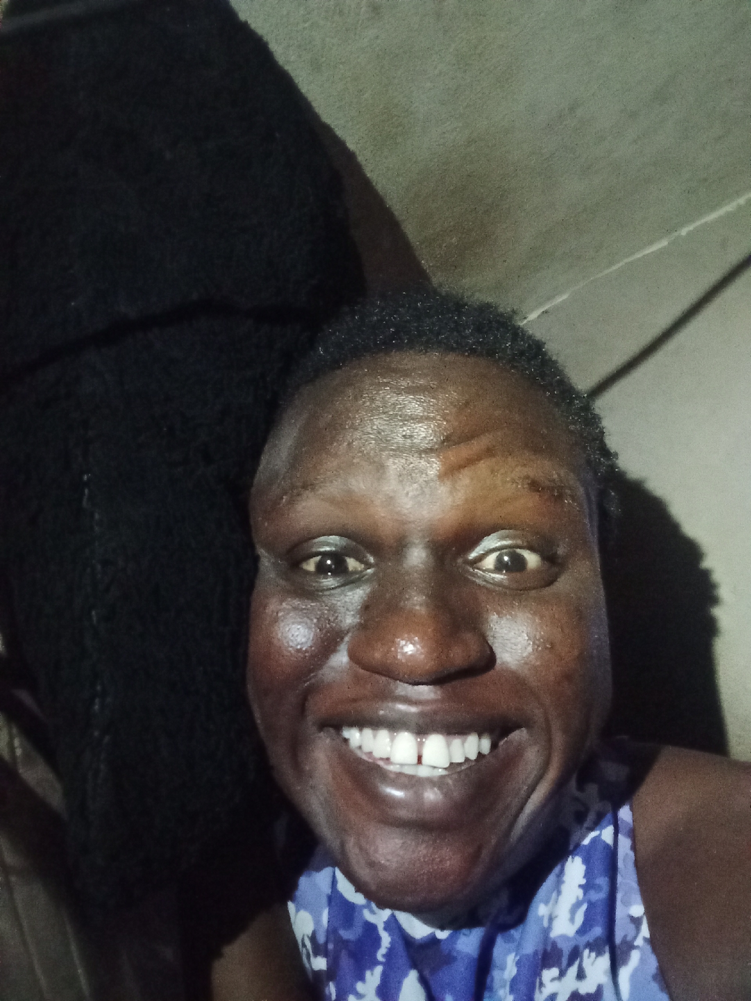 A close-up indoor shot shows a smiling dark-skinned person wearing a blue and white patterned sleeveless shirt. The person has short black hair and prominent teeth. A dark object is visible to the left of the person. The background is a plain, light-colored wall.
Location: Unknown, Unknown.