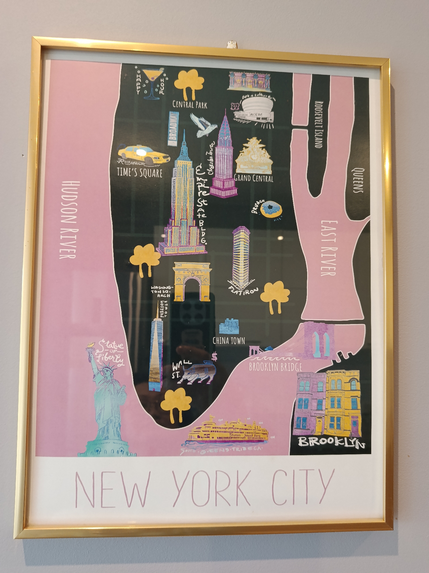 The image shows a framed illustration of a stylized map of New York City. The Hudson River is marked on the left side and the East River is to the right. Key locations and landmarks are depicted, including the Statue of Liberty, Wall Street, Chinatown, Brooklyn Bridge, Brooklyn, the Flatiron Building, Washington Square Arch, Grand Central Terminal, the Chrysler Building, the Empire State Building, Times Square, Central Park, Roosevelt Island, and Queens. The text "NEW YORK CITY" is printed at the bottom. The illustration has a black background with pink areas denoting the rivers and some landmasses. It is framed in a gold-colored frame.
There is no indication of the location Odintsovo, Russia in the image.