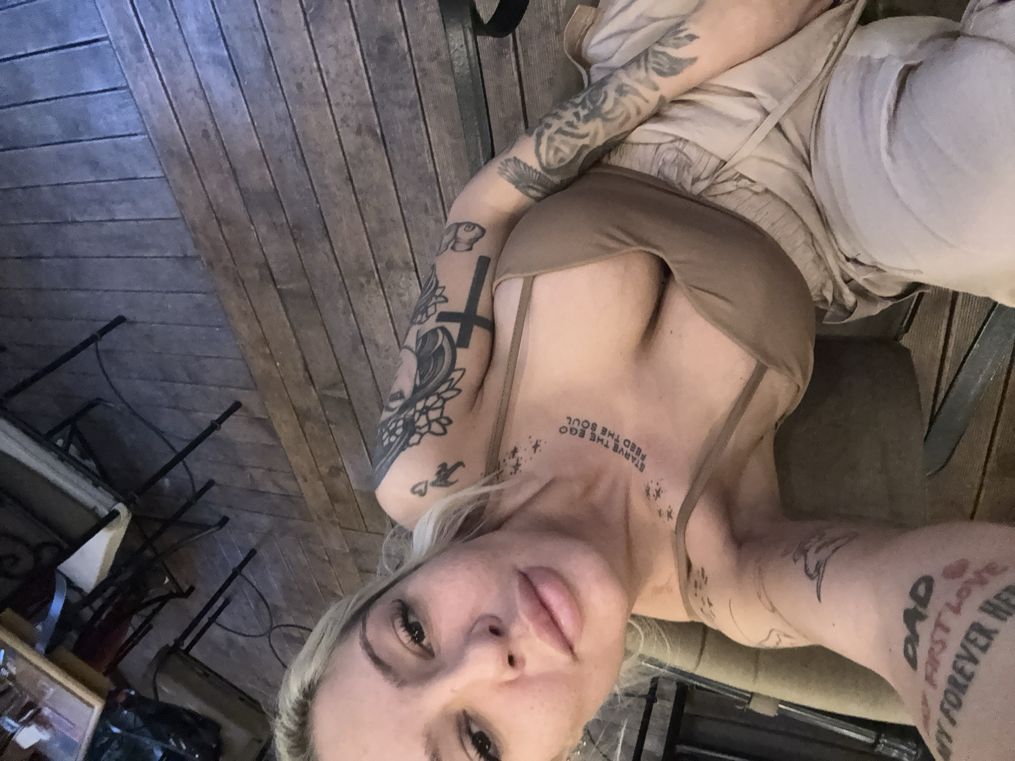 A fair-skinned person with blonde hair and multiple tattoos is lying on what appears to be a wooden deck or floor. They are wearing a brown top and beige bottoms. Tattoos are visible on their arms, neck, and chest, including a cross, floral designs, text ("STARVE THE EGO FEED THE SOUL"), and a "DAD" tattoo. In the background, there is furniture, possibly chairs, and wooden planks. The image is taken from a close-up perspective, looking down at the person. There is no indication of the location in the city of Plovdiv, Bulgaria.
