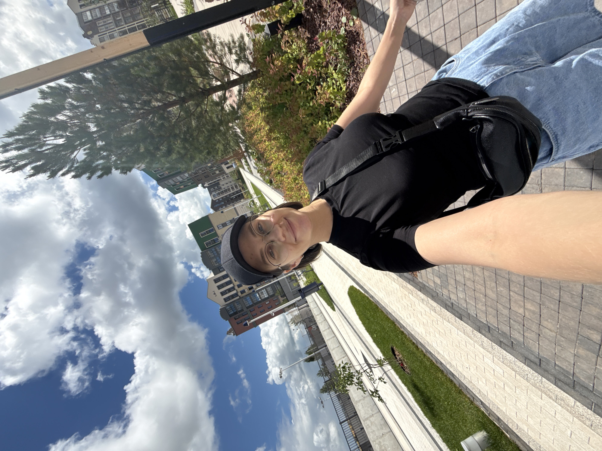 In Izhevsk, Russia, a young woman with dark hair, glasses, and a black baseball cap takes a selfie on a sunny day. She is wearing a black t-shirt, blue denim shorts, and a black crossbody bag. The background features a modern urban landscape with colorful apartment buildings, a paved walkway, lush green grass, and a tall pine tree. The sky is bright blue with scattered white clouds.