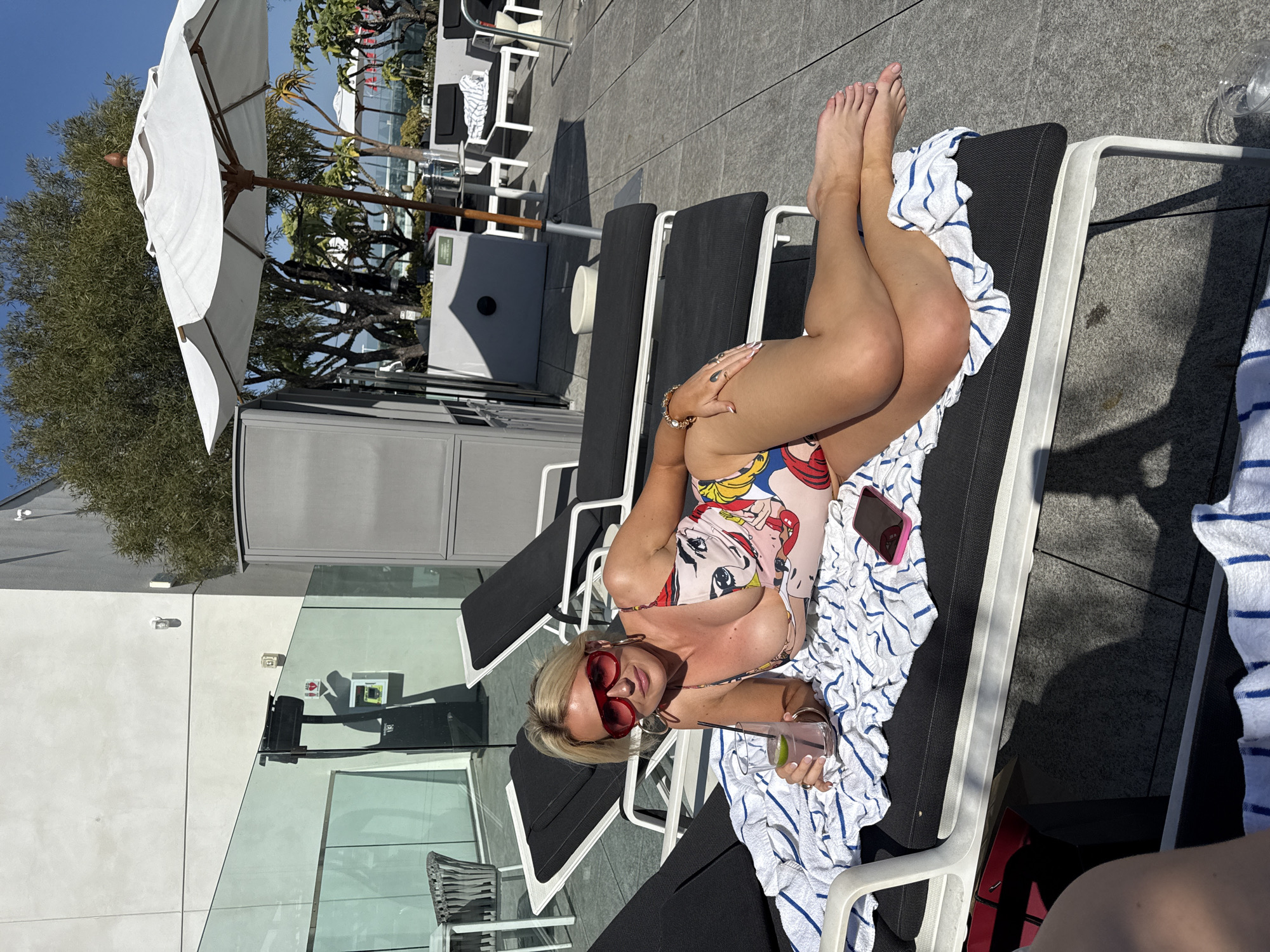 A woman is lying on a black lounge chair on a sunny day. She is wearing a comic-print swimsuit and red heart-shaped sunglasses. Her blonde hair is styled short. She is holding a drink in her right hand. The lounge chair has a white towel with blue stripes. A smartphone is visible near her. Behind her are more lounge chairs and a white umbrella. In the background are buildings, suggesting an urban setting. The location is city of Unknown, Unknown.