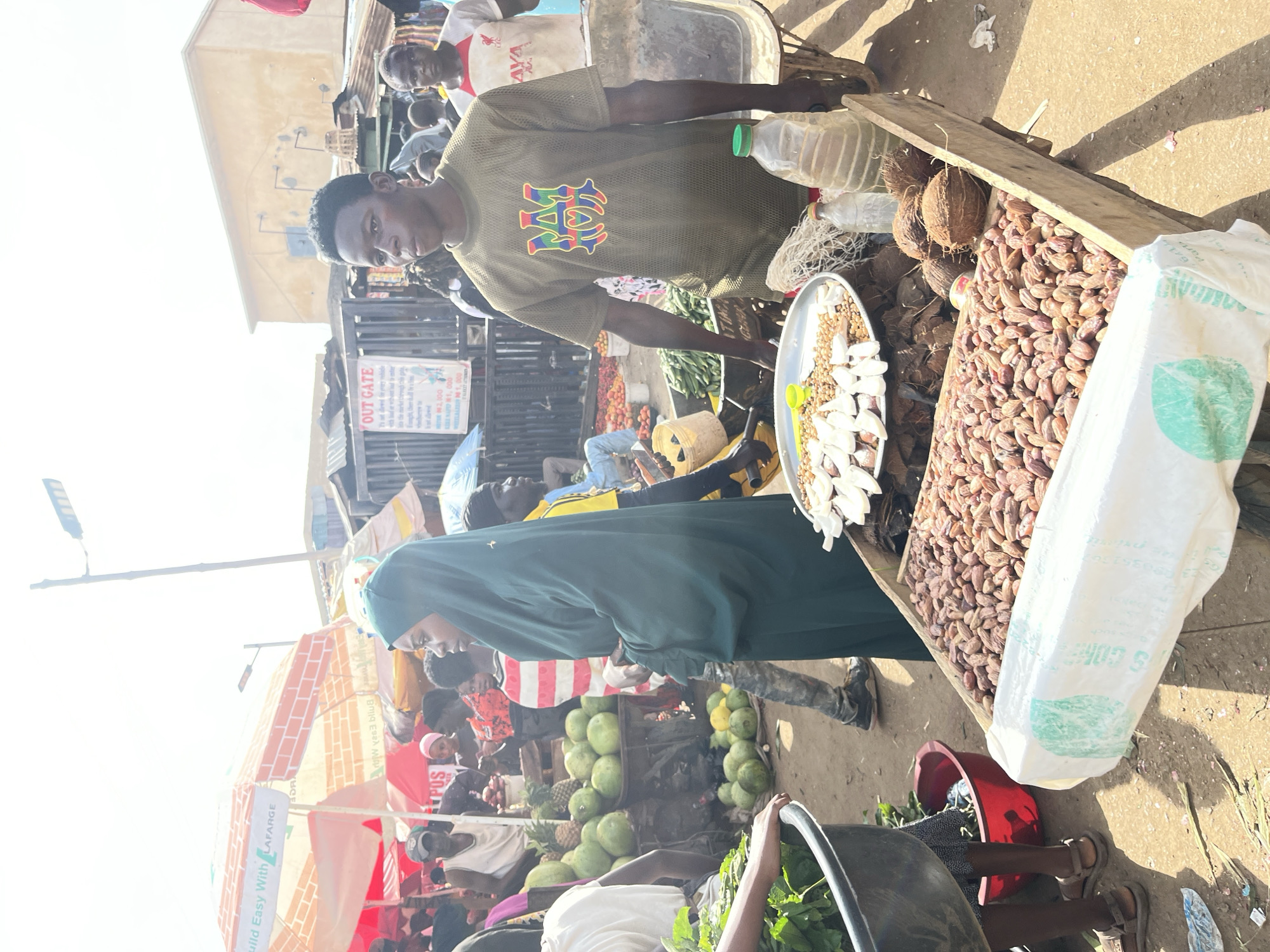 In Rafin Par, Nigeria, a bustling outdoor market scene unfolds. The image captures a vibrant marketplace with various stalls and vendors. In the foreground, a wooden cart is laden with goods, including what appears to be dried dates and coconuts. To the left, a person's hands are seen reaching into a large black basin filled with leafy greens, suggesting fresh produce is being handled. Behind this, a red bucket is visible.
A young woman, draped in a dark green hijab and dress, stands prominently, her gaze directed slightly away from the camera. Beside her, a young man wearing a khaki mesh t-shirt with a colorful emblem, stands looking forward. Further back, a man in a white t-shirt with a red Liverpool logo is also visible. The market is filled with other people, some blurred in the background, indicating a lively atmosphere.
Overhead, umbrellas provide shade, with one prominently displaying the "LAFARGE" logo, suggesting a commercial presence in the area. In the distance, a gate with an "OUT GATE" sign is visible, and the urban environment is characterized by low-rise buildings and a clear sky. The ground appears to be dusty, typical of an outdoor market in a dry climate. The overall impression is one of active commerce and daily life in a Nigerian city.