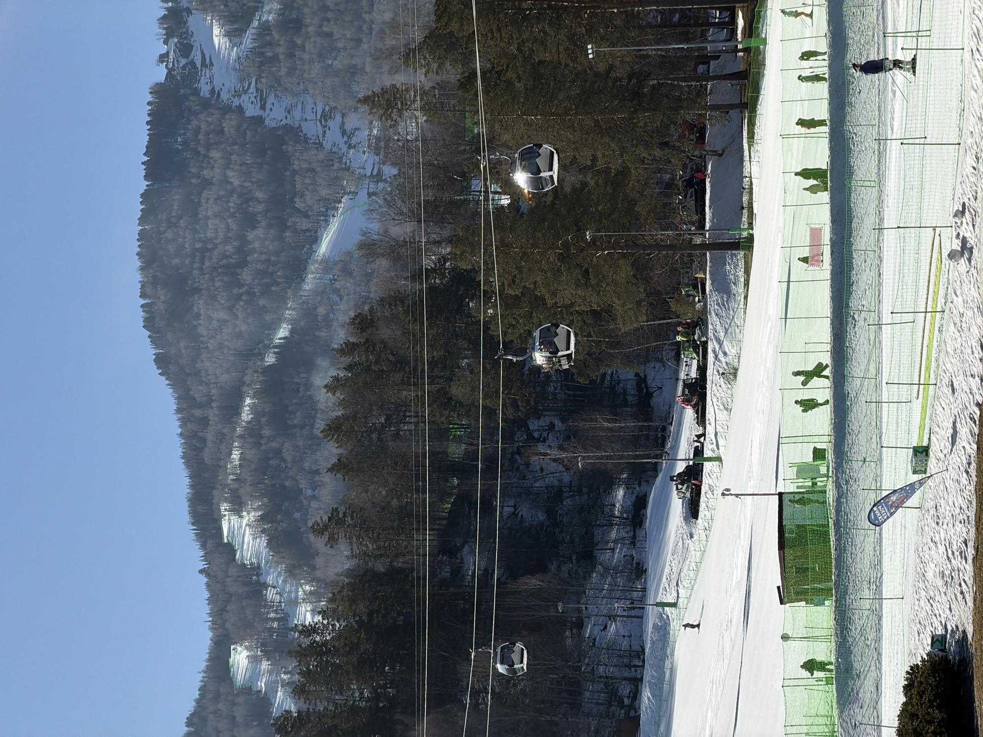 The image shows a ski resort in Озёрное, Russia, on a sunny day. A ski lift with several gondolas is visible, ascending the wooded mountain. The ski slope is covered in snow, with a green safety net running along its side, and several skiers are present. In the foreground, there is a snow-covered area with a partially visible blue and white flag.