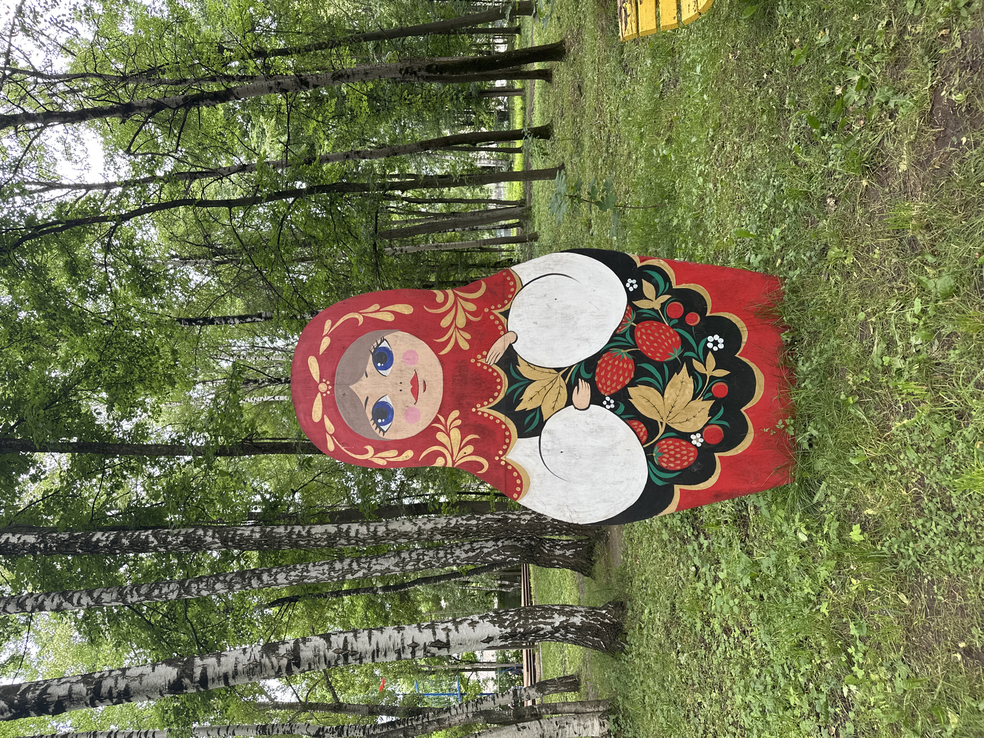 A large, wooden Matryoshka doll, painted in traditional Russian style, stands in a park in Izhevsk, Russia. The doll is red with gold floral patterns and a depiction of strawberries. It is surrounded by green grass and trees, with a yellow bench visible to the right. The background shows a playground structure and more trees.