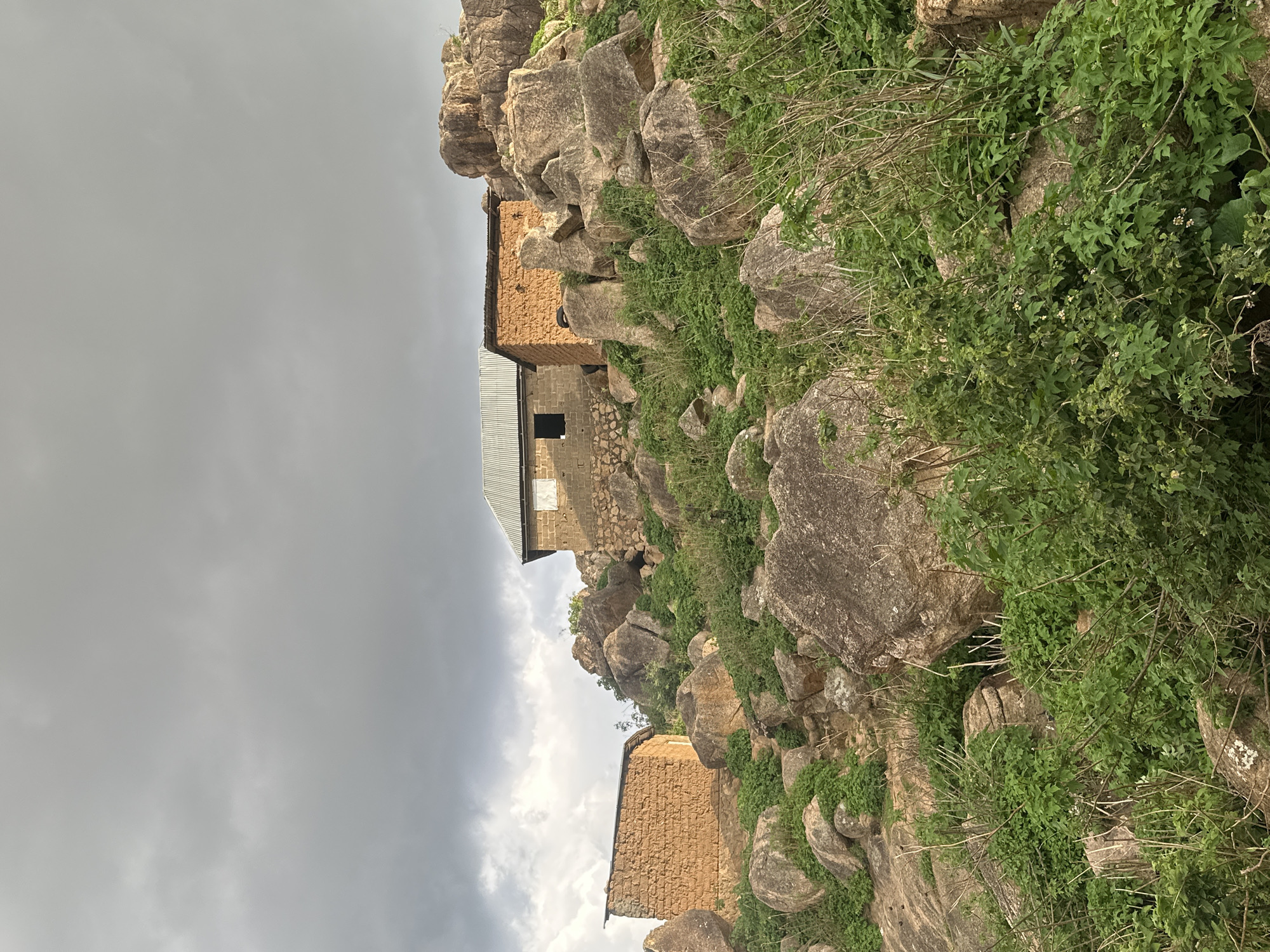 A panoramic view reveals a rugged, rocky landscape under a heavily overcast sky. The image depicts a community built amidst large, weathered rock formations that dominate the terrain. Lush green vegetation, including bushes and smaller plants, extensively covers and grows between the rocks, suggesting a fertile environment despite the rocky ground.
Several modest structures are visible, skillfully integrated into the natural environment. In the mid-ground, a prominent building features walls constructed from earth-toned materials, possibly mud bricks or adobe, topped with a corrugated metal roof. A dark, rectangular opening on its facade could be a window or doorway. Further down the rocky incline and to the left, another building with similar earth-toned walls and a corrugated metal roof is partially visible. Higher up and to the right of the central structure, a third building stands, characterized by concrete block walls and a corrugated metal roof.
The sky is a uniform, heavy gray, indicating a cloudy day with diffused light, possibly before or after rainfall. No people are present, and no specific activities are taking place, lending a quiet and serene atmosphere to the scene. The architecture and natural setting are indicative of a rural or semi-urban area, consistent with the provided location context of Jos, Nigeria, which is known for its distinctive rock formations and hilly terrain. There is no visible text.