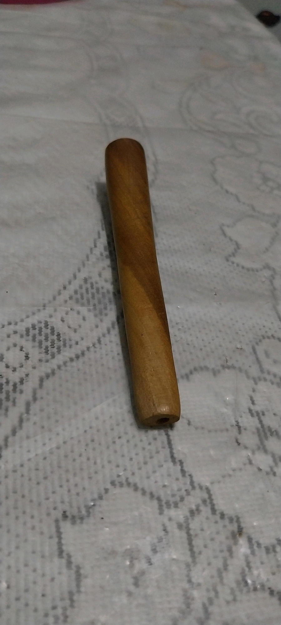 A single, short, cylindrical wooden object with a hole at one end rests on a light-colored, patterned fabric. The object is oriented vertically and appears to be made of light brown wood with visible grain. The fabric beneath has a repeating, raised pattern that resembles lace or embroidery. The lighting suggests an indoor setting, with shadows cast on the fabric. The specific location of Bojong Nangka, Indonesia, is not depicted in the image itself.