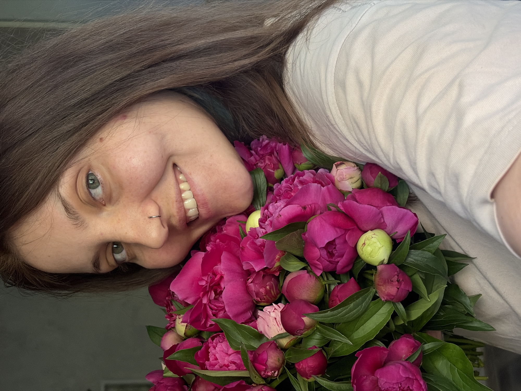 A close-up, medium shot shows a woman with brown hair and green eyes, smiling at the camera, holding a large bouquet of bright pink peonies. She is wearing a light-colored t-shirt. The background is indistinct and appears to be an indoor setting, possibly a home. The image does not provide any specific information about its location in Chaikovsky, Russia.