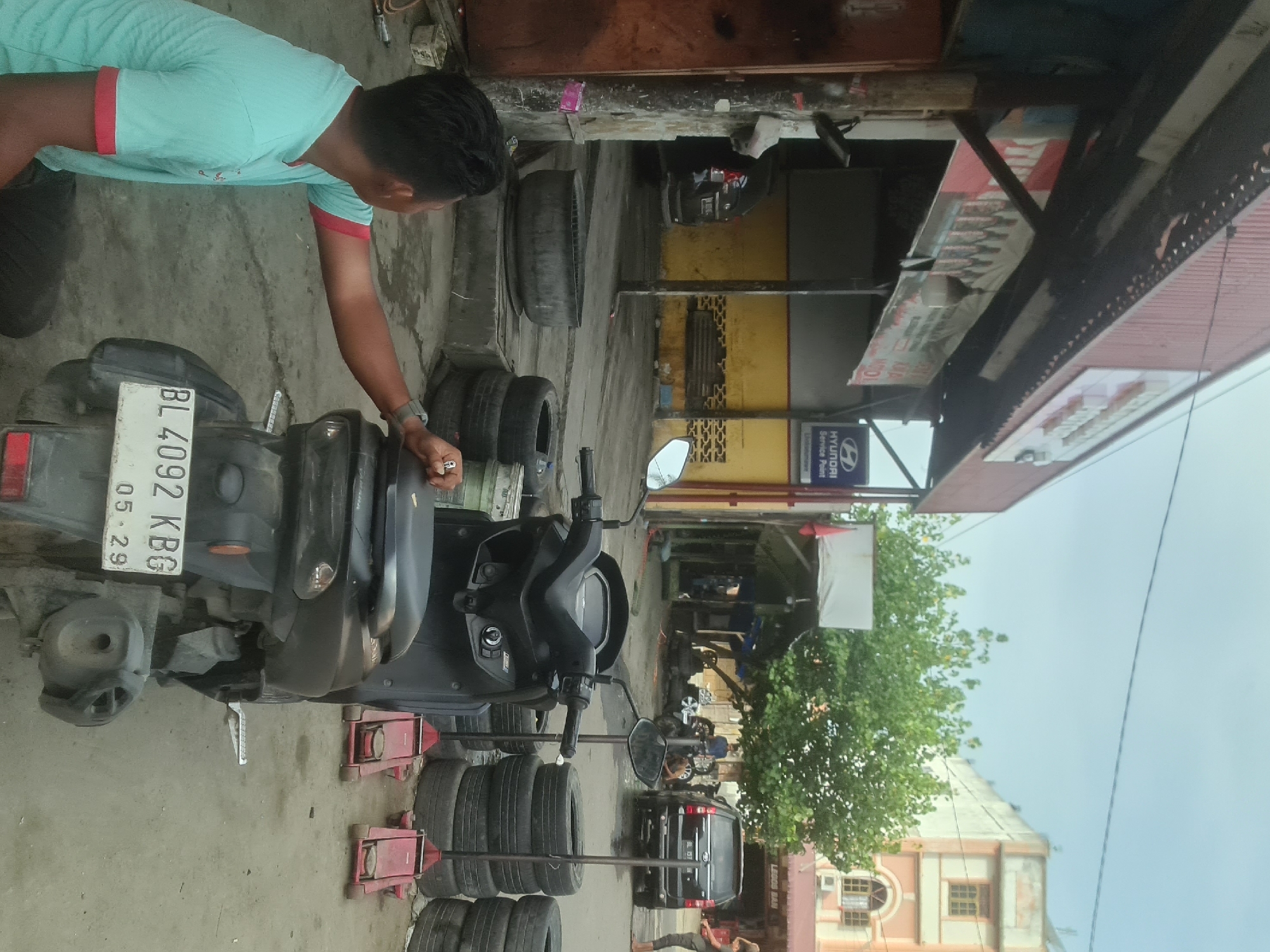 The image captures an outdoor scene at what appears to be a motorcycle repair shop or tire service center in Lhokseumawe, Indonesia. In the foreground, a person wearing a light blue t-shirt with red trim is kneeling down, touching the back of a black scooter. The scooter has a license plate that reads "BL 4092 KBG 05-29". Behind the scooter, several stacks of tires are visible, along with two red hydraulic jacks.
The background reveals a covered area with a red roof, likely part of the shop. A sign with a Hyundai logo and "Service Point" is mounted on the building. Further back, a black SUV is parked. There are other motorcycles and a few people milling about in the distance. Trees and a multi-story building with an arched window are visible behind the shop. The ground is paved with concrete, and the overall impression is of a functional, working environment.