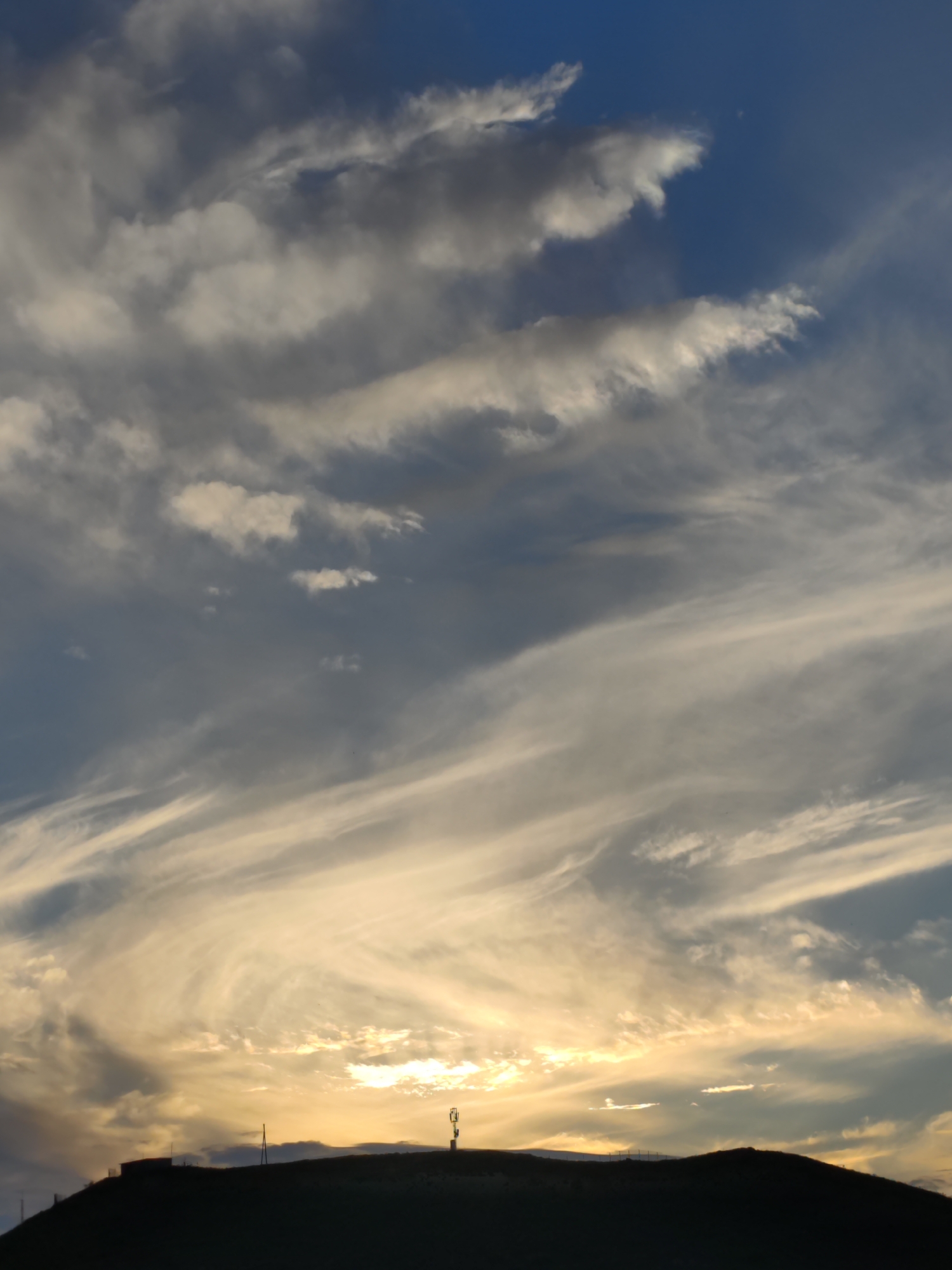 A silhouette of a rounded hill dominates the lower portion of the image, with the dark outline of structures and a cell tower visible against a bright horizon. The sky is filled with a dramatic display of clouds, ranging from wispy, light-colored cirrus clouds that catch the golden light of sunset, to darker, more opaque clouds that create depth and contrast. The overall impression is of a serene, natural landscape at dusk. The location is not discernible from the image itself, and there are no people or animals present.