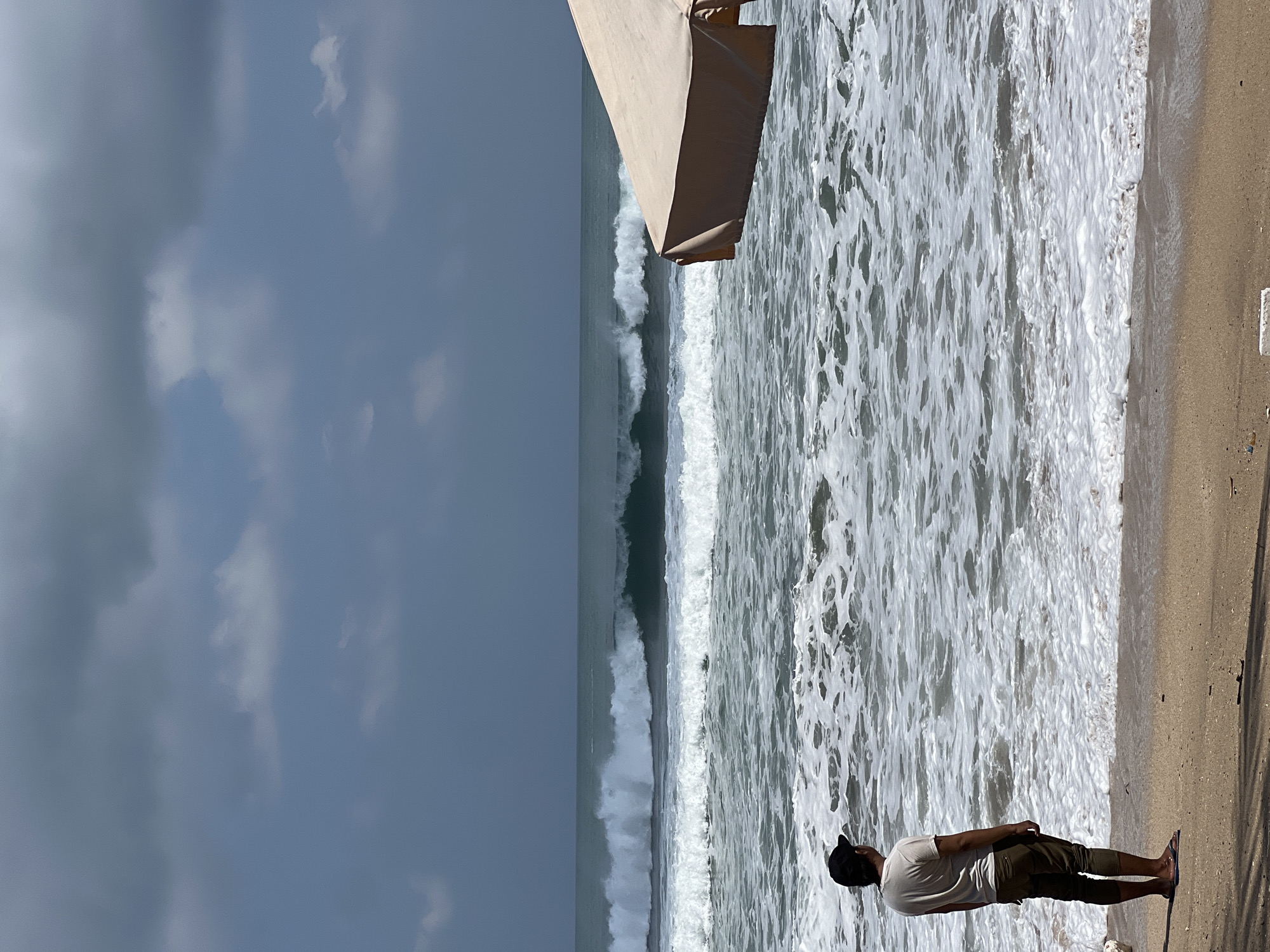 A man stands on a sandy beach in Kuta, Indonesia, facing the ocean. He is wearing a light-colored t-shirt, dark pants, and a black cap. His back is to the camera, and he is looking out at the waves. The ocean is rough, with large waves breaking on the shore. The sky is overcast with dark clouds, and there is a beach umbrella in the foreground on the right side of the image. The sand is wet and dark near the water's edge, transitioning to a lighter, drier sand further up the beach. Debris, including plastic, is scattered on the sand.