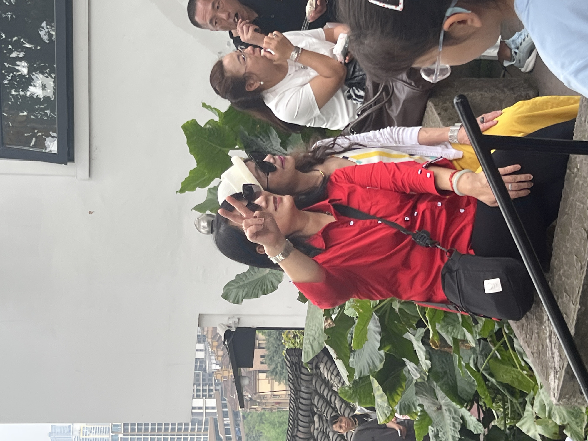 The image shows a group of people posing for a photo. In the foreground, a woman with dark hair wearing a red shirt and black bag is holding up a peace sign. Behind her, another woman wearing sunglasses and a white head covering is leaning back. To the left is a white wall with a window. In the background, there is a cityscape with various buildings and greenery. The location is in the city of Nan'an District, China.