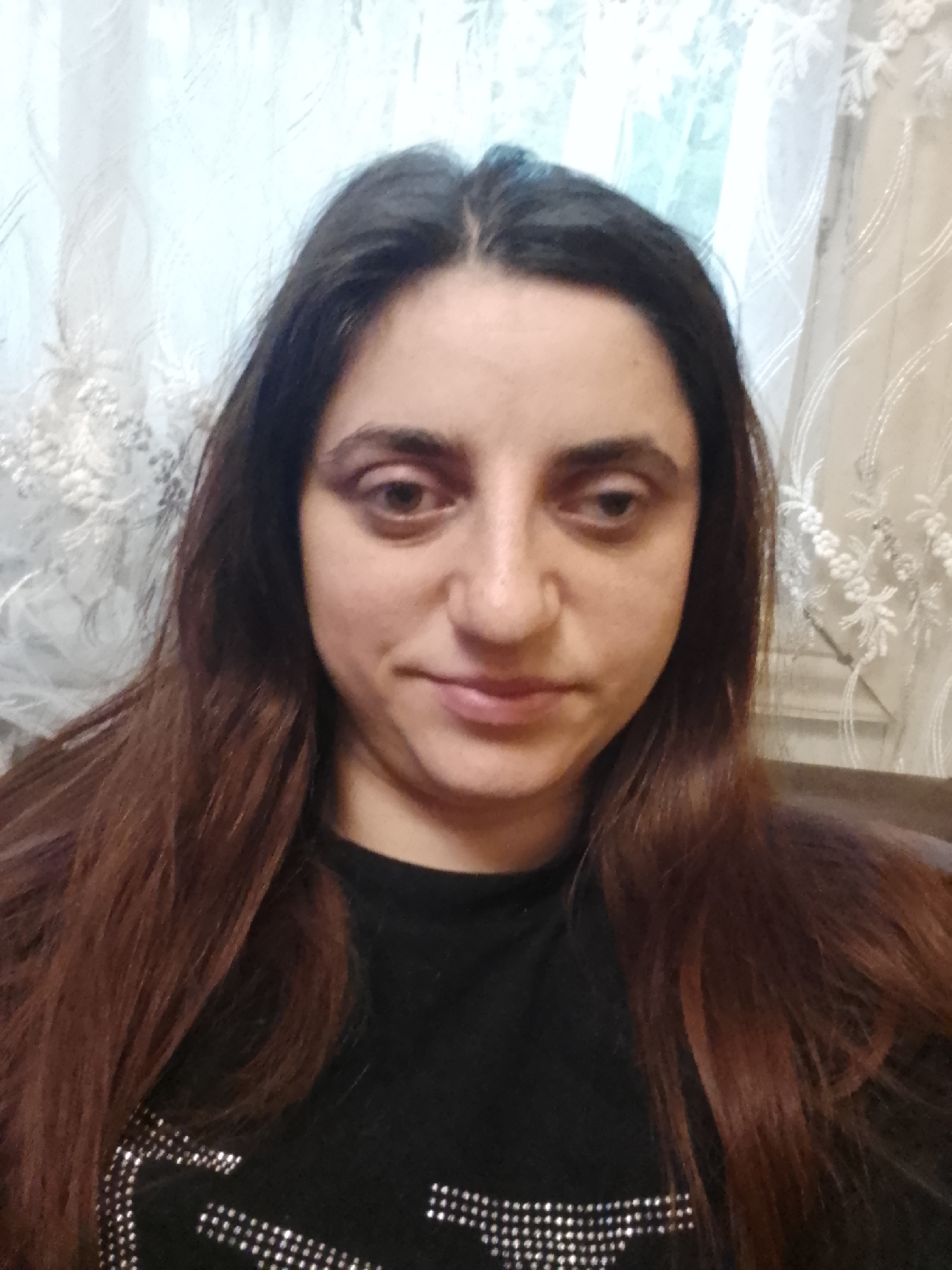 A woman is depicted in a head-and-shoulders portrait, positioned centrally in an indoor setting in Yerevan, Armenia. She has long, dark brown hair, parted in the middle, extending over her shoulders. Her eyes are brown, with the right eye (viewer's left) appearing partially closed, while the left eye (viewer's right) is more open. Her facial expression is subtle, with closed lips hinting at a slight smile. She is wearing a black garment, possibly a top or sweater, which features silver-colored studded or rhinestone embellishments arranged in patterns across her chest and right shoulder. The background consists of light-colored, possibly white or cream-colored, sheer curtains or drapes adorned with intricate white lace-like floral or geometric patterns. No other subjects or significant objects are visible, and no specific action is depicted beyond the pose for the portrait.