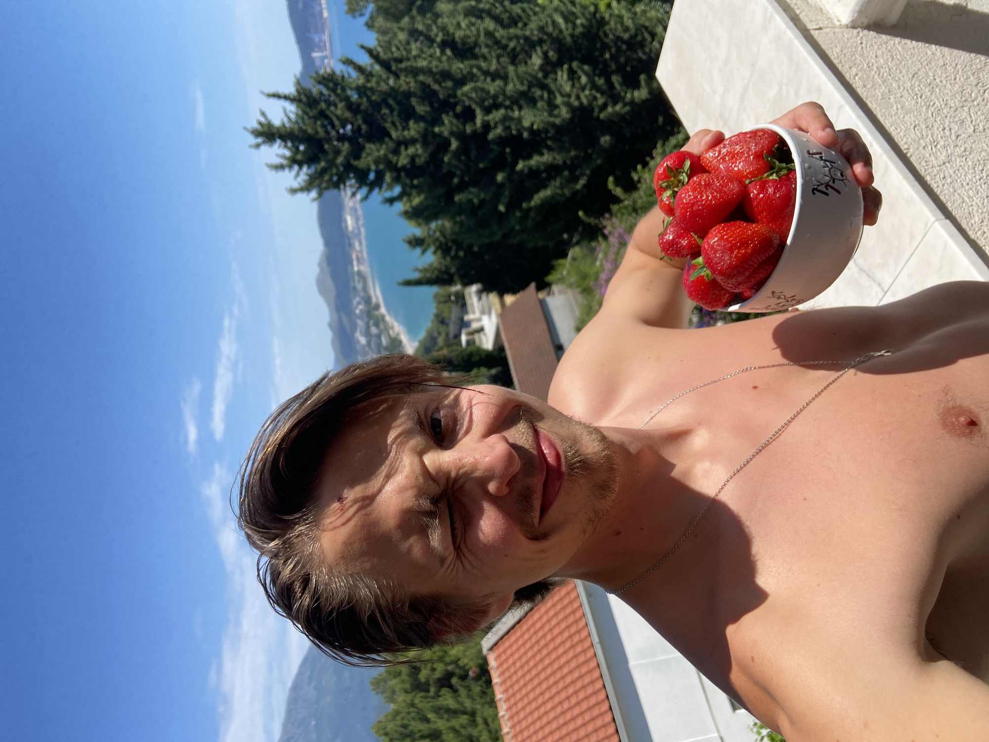 A shirtless man with short brown hair and a silver necklace holds a white bowl filled with red strawberries in his right hand. The photograph is taken from a low angle, showcasing his face and the bowl. He has one eye squinted shut. In the background, there is a view of the sea, trees, and buildings, indicating an elevated location. The location is in the city of Šušanj, Montenegro, as suggested by the Mediterranean landscape and architecture in the background.