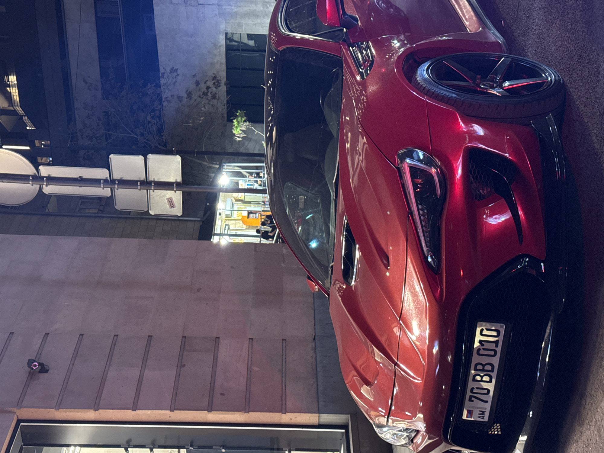 A glossy red sports car, likely a Toyota 86 or Subaru BRZ, is prominently featured in an urban street scene at night or late evening. The car is parked, showcasing its sleek design, pronounced headlights, and a large black mesh grille on the front bumper. It is equipped with black multi-spoke wheels, which have subtle red accents.
The vehicle's license plate, clearly visible on the front bumper, reads "AM 70 BB 010," indicating its origin from Armenia.
In the background, to the left of the car, a street pole supports multiple white rectangular and one circular sign, suggesting traffic or informational markers. Beyond these, dark buildings with illuminated windows and a tree with bare branches are visible under the artificial lighting of the city. To the right and behind the car, a building facade with a textured, possibly concrete or stone surface and vertical grooves dominates the background. The scene is illuminated by various artificial light sources, creating reflections on the car's metallic red paint and highlighting the surrounding urban environment, consistent with a street in Yerevan, Armenia.