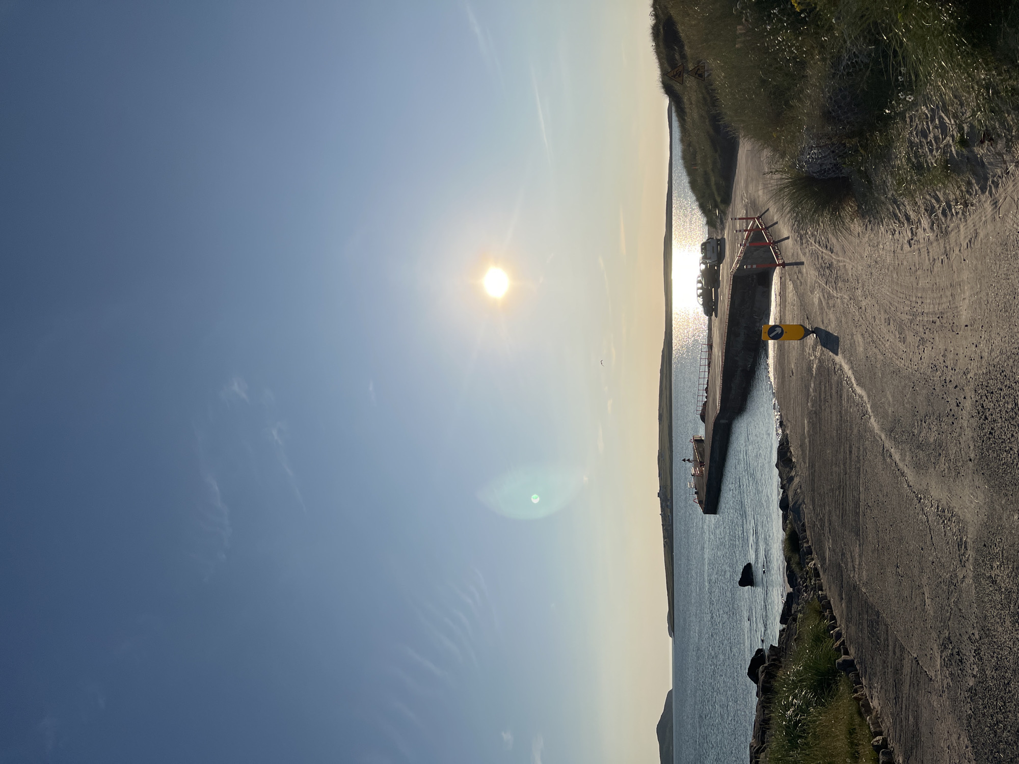 This is a photograph taken in Glassagh Lower, Ireland, depicting a coastal scene at sunset. The sun is visible in the upper right quadrant of the image, casting a bright, warm glow and its reflection on the water. A concrete pier extends into the calm sea from the right side of the frame, with two cars parked near its base. A short concrete ramp leads down to the water's edge in the foreground, with sandy terrain and grassy dunes flanking it on the right. Rocks and some sparse vegetation are visible along the shoreline. The sky is a clear blue with a few wispy clouds, and the overall atmosphere is serene and natural.