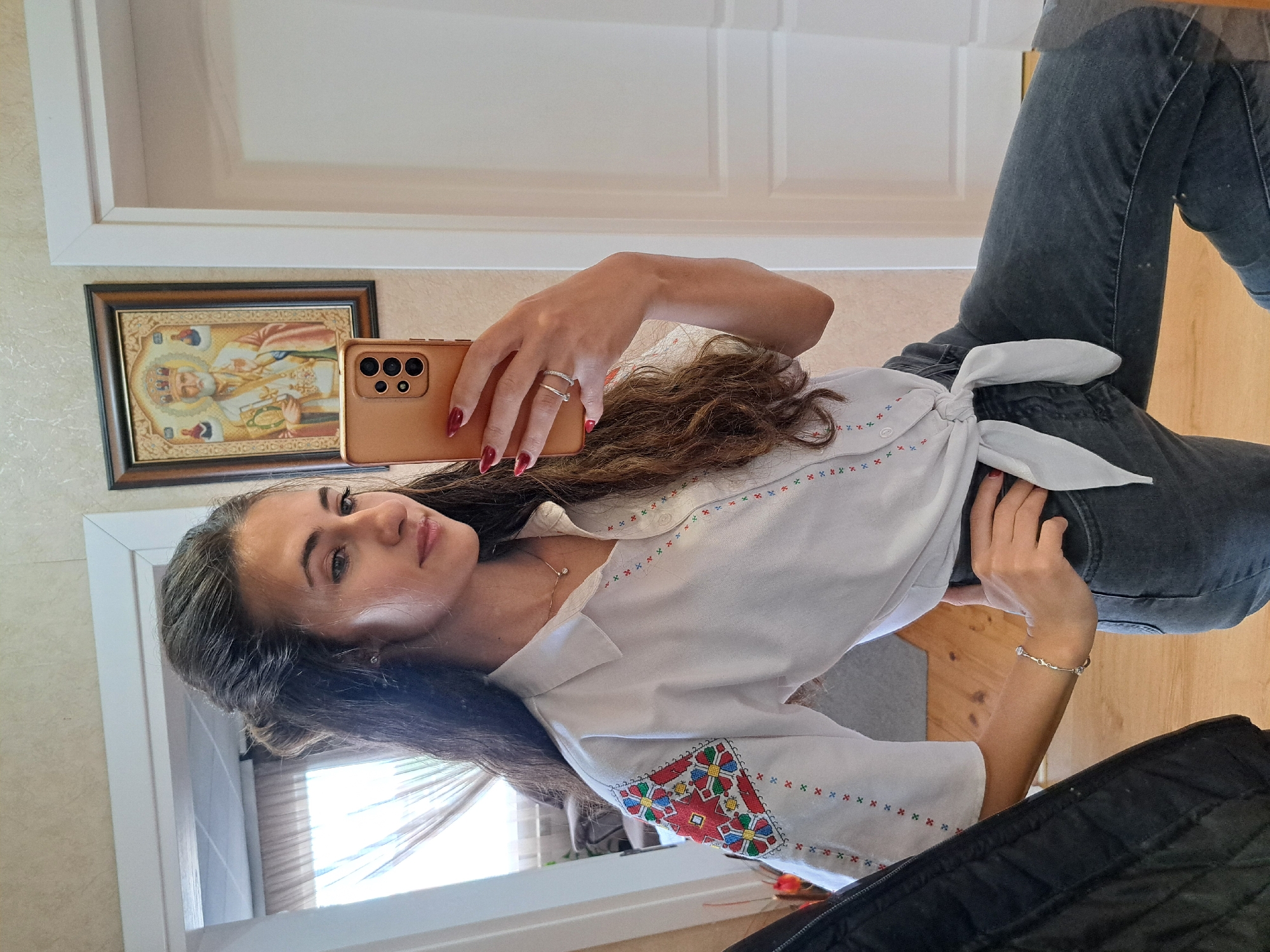 A woman is shown in a selfie shot taken indoors. The room seems to be a house or apartment, with visible walls, a wooden floor, and details like a door frame and a framed icon hanging on the wall. The woman is holding a smartphone to take the picture. She is wearing a white shirt with embroidered details and dark jeans tied at the waist with a white belt. Her hair is long and wavy. She is wearing jewelry, including rings on her fingers and a bracelet on her wrist. The lighting in the room appears natural, possibly from a window. The image is taken at a slight angle, with the woman looking at the camera.