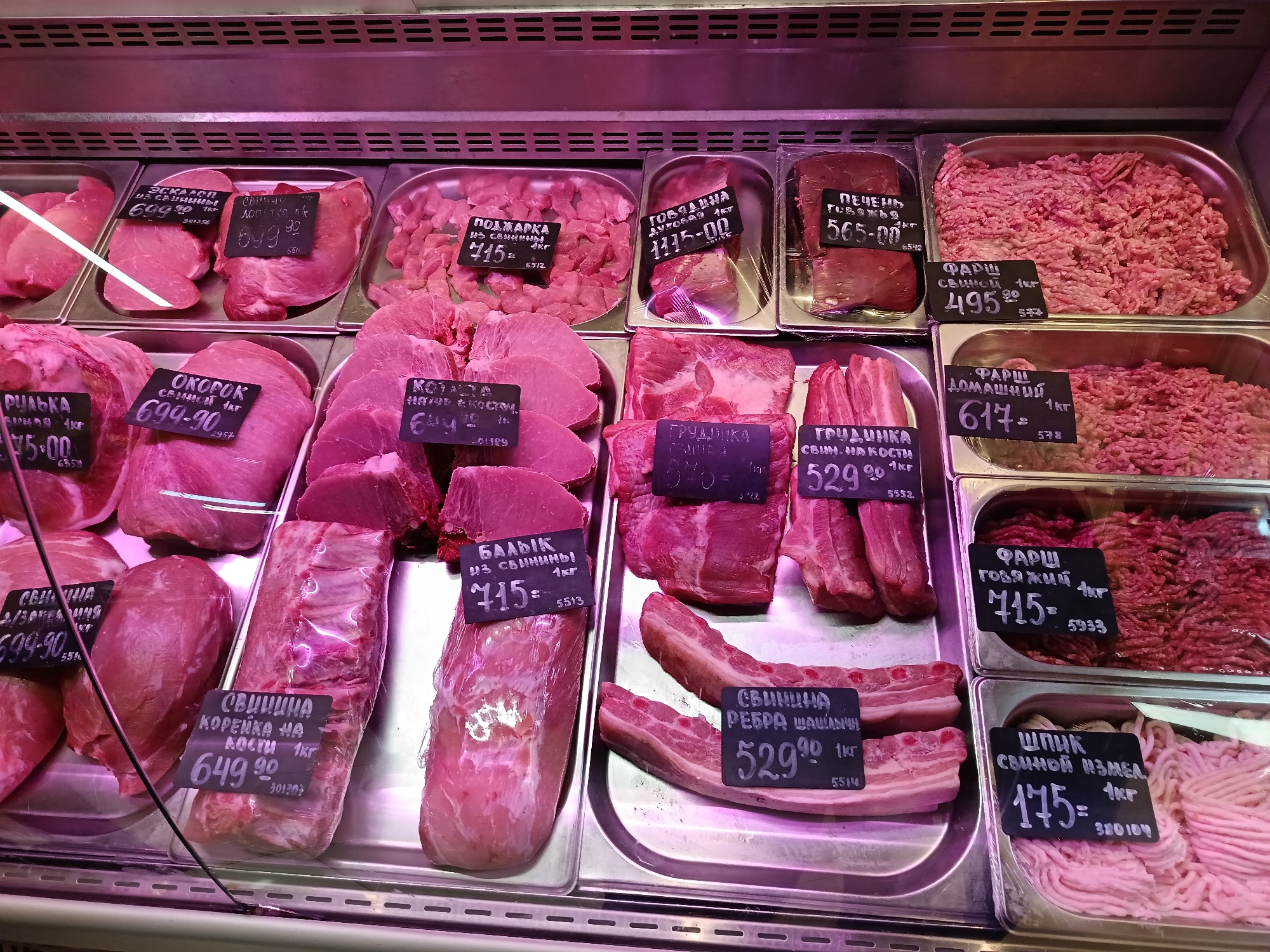 This is a photograph of a refrigerated meat display case. The display is well-stocked with various cuts of raw meats, including pork, beef, and ground meat. Each cut of meat is placed on a metal tray and accompanied by a price tag written in Cyrillic script. The labels include information such as the type of meat, its cut, and the price per kilogram. The arrangement of the meat in the display case suggests a supermarket or butcher shop setting. Given that the OCR output indicates the meat prices in Russian currency, and the label texts are in Russian, it is likely this scene is set in Russia, and possibly as indicated by the prompt, the city of Osa.