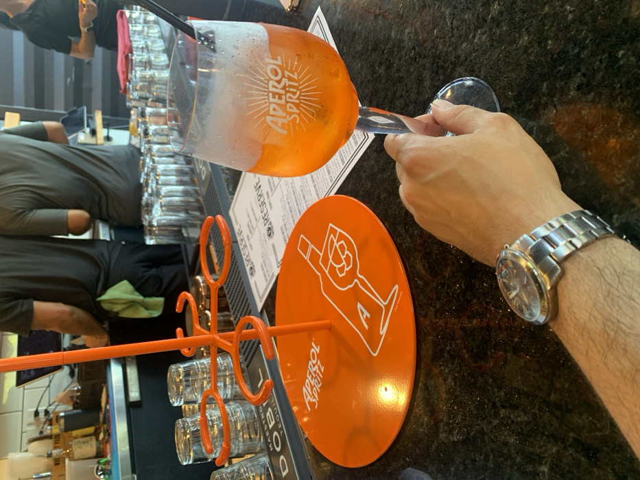 A close-up shows a hand holding a large Aperol Spritz glass filled with the orange drink and ice. The glass displays the Aperol Spritz logo. Beside it sits an orange Aperol Spritz branded coaster with a white outline of a bottle and glass. A watch is worn on the wrist of the person holding the glass. A bartender is working at the bar. Behind the glass stand a menu and an orange glass holder containing several small glasses. The countertop is dark and speckled.