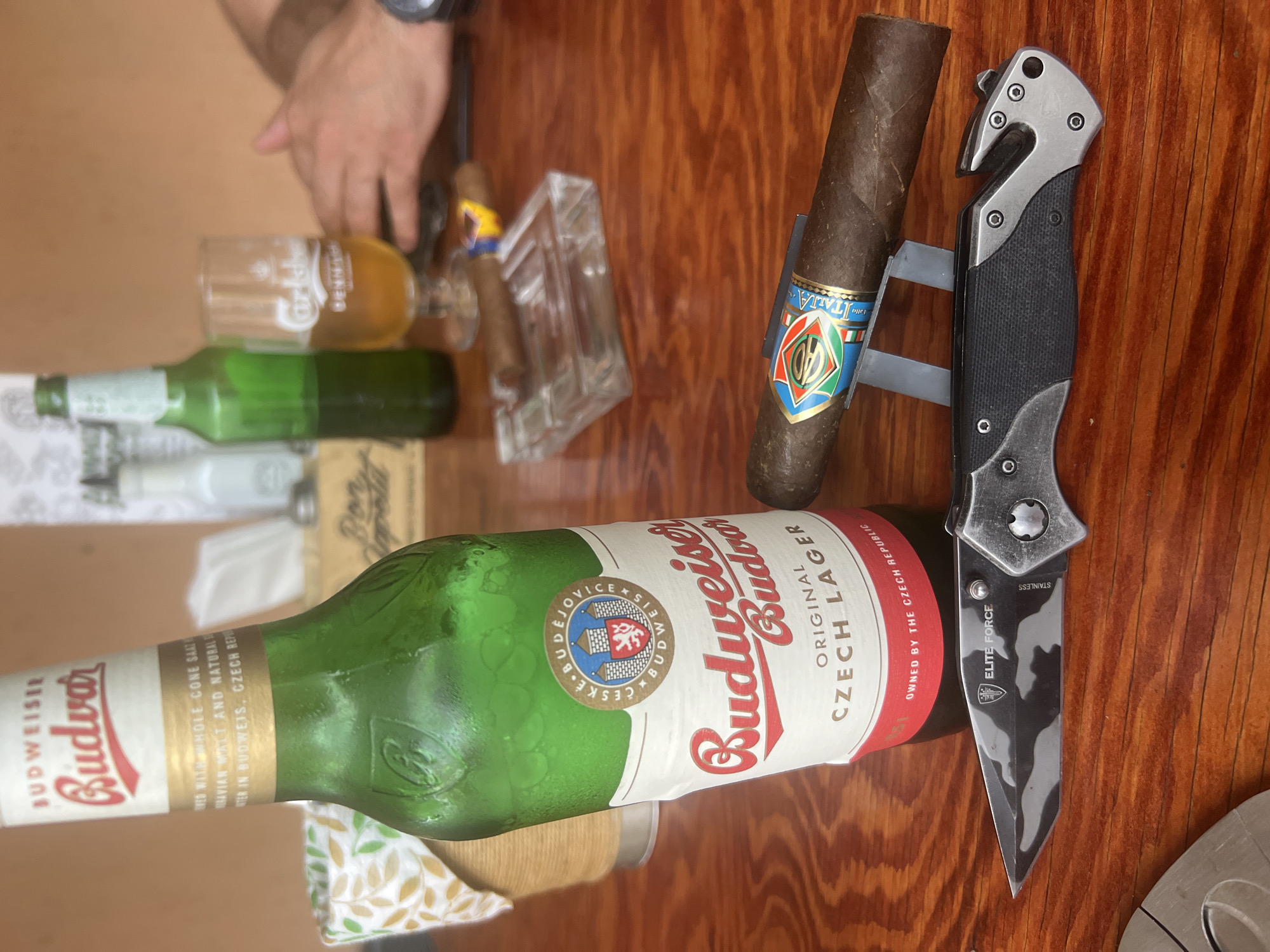 This image captures a close-up, indoor scene on a polished reddish-brown wooden table, depicting a casual leisure setting within a bar or restaurant environment in Sofia, Bulgaria.
The central focus is a chilled, green glass bottle of Budweiser Budvar Original Czech Lager, displaying prominent condensation. Its label features "BUDWEISER Budvar," "ČESKÉ BUDĚJOVICE," and "OWNED BY THE CZECH REPUBLIC." To the right of the bottle, a dark brown cigar is horizontally positioned on a small, grey metal stand. The cigar's colorful band prominently displays the letters "PH" and the word "ITALIA." In the immediate foreground, a partially open Elite Force folding knife rests on the table. The knife features a black and grey camouflage-patterned tanto-style blade marked with the Elite Force emblem and "STAINLESS." Its handle is black textured with silver metallic components, and it incorporates a distinct seatbelt cutter/glass breaker tool at the pommel. A circular metallic object is partially visible in the bottom-left corner of the frame.
In the midground, a clear glass ashtray contains a partially smoked cigar and a colorful wrapper. Behind the ashtray, a Carlsberg-branded glass holds a small quantity of amber liquid. Another out-of-focus green bottle is visible behind the Carlsberg glass.
The background is characterized by a blurred, plain light brown wall. A white napkin dispenser with a green and yellow leaf pattern is visible on the left. A box labeled "Bon Apetita" is situated behind the beer bottle, alongside several other out-of-focus condiment bottles and shakers. To the upper right, the forearms and hands of an individual are visible, resting on the table, with a dark-colored watch on the left wrist. The arrangement of objects suggests a moment of relaxation, likely involving beverages and smoking.