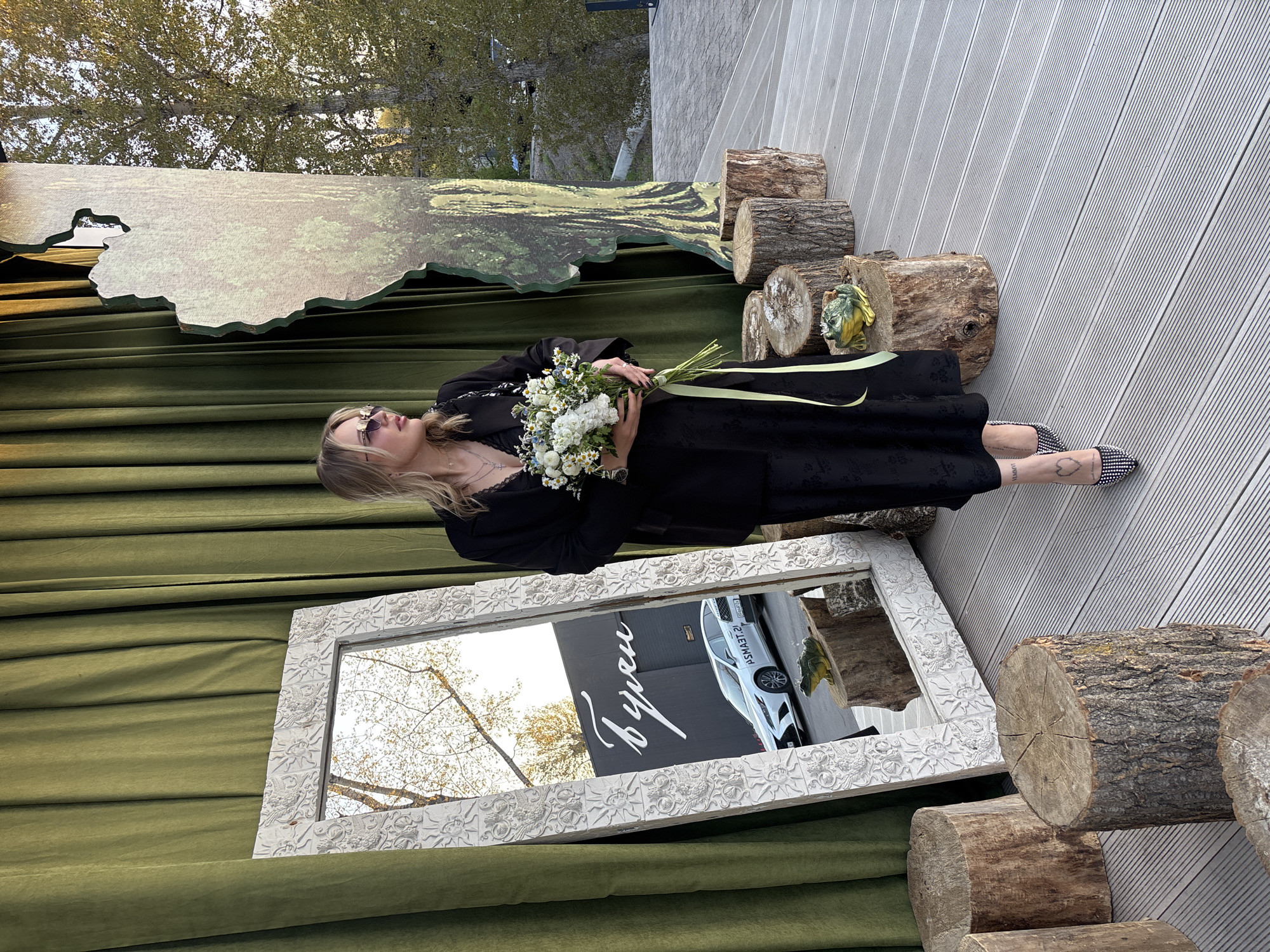 A photograph captured in Krasnoyarsk, Russia, depicts a woman in a black dress holding a bouquet of flowers, positioned in front of a mirror. The mirror reflects a white car parked near a building adorned with the word "Byron" in Cyrillic script. The woman is also wearing sunglasses and patterned shoes. The setting includes green draped fabric, wooden logs, and a cut-out backdrop resembling a forest scene. The outdoor scene suggests an urban environment with natural elements incorporated into a staged or decorated space.