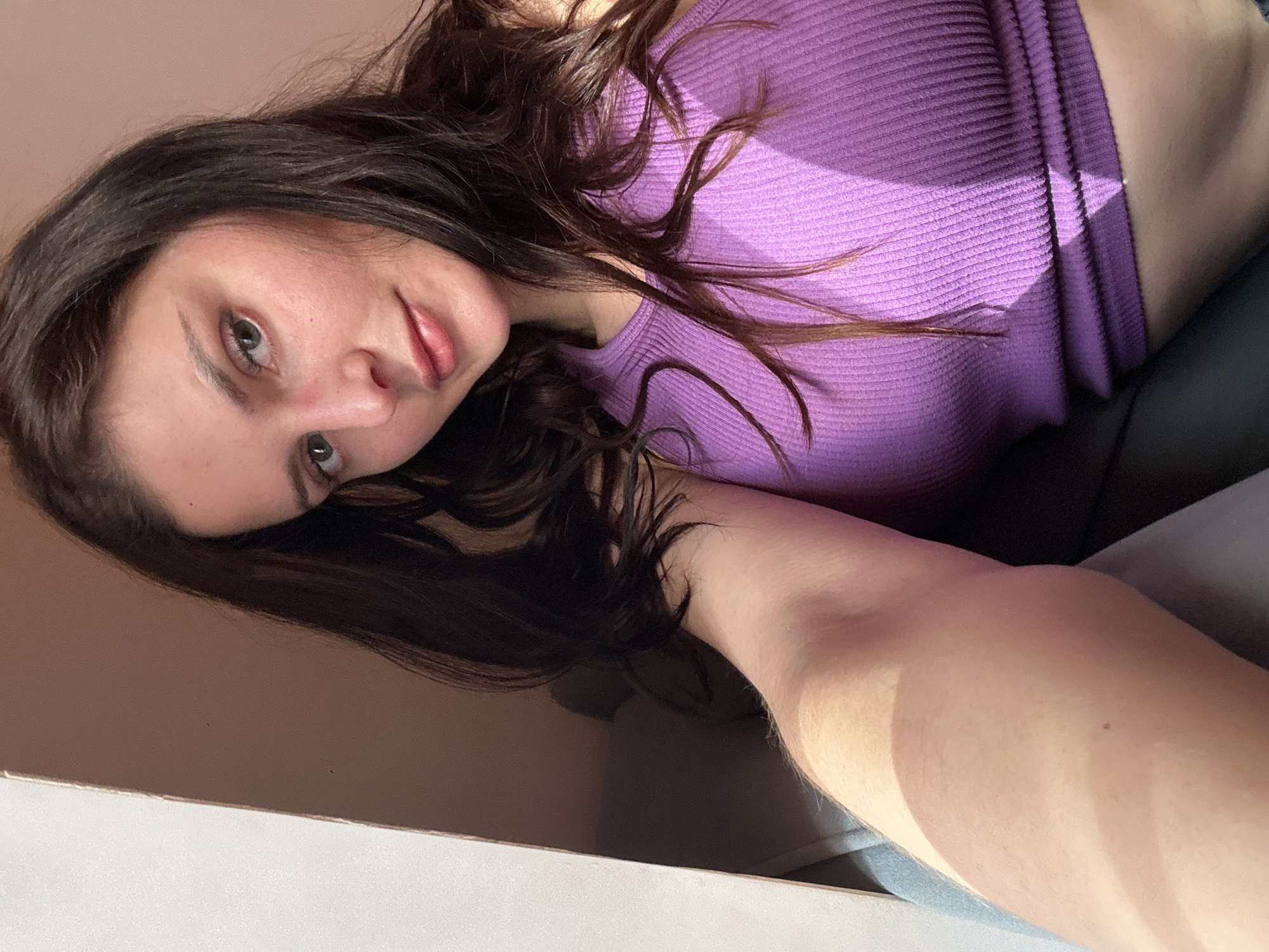 This is a close-up, sideways selfie of a young woman with long, dark, wavy hair. She is wearing a purple, ribbed, sleeveless top. Her left arm is visible, resting against what appears to be a dark surface. She is looking towards the camera with a neutral expression. The lighting suggests it is daytime, with sunlight casting shadows across her top and arm. The background is a plain, light brown wall.