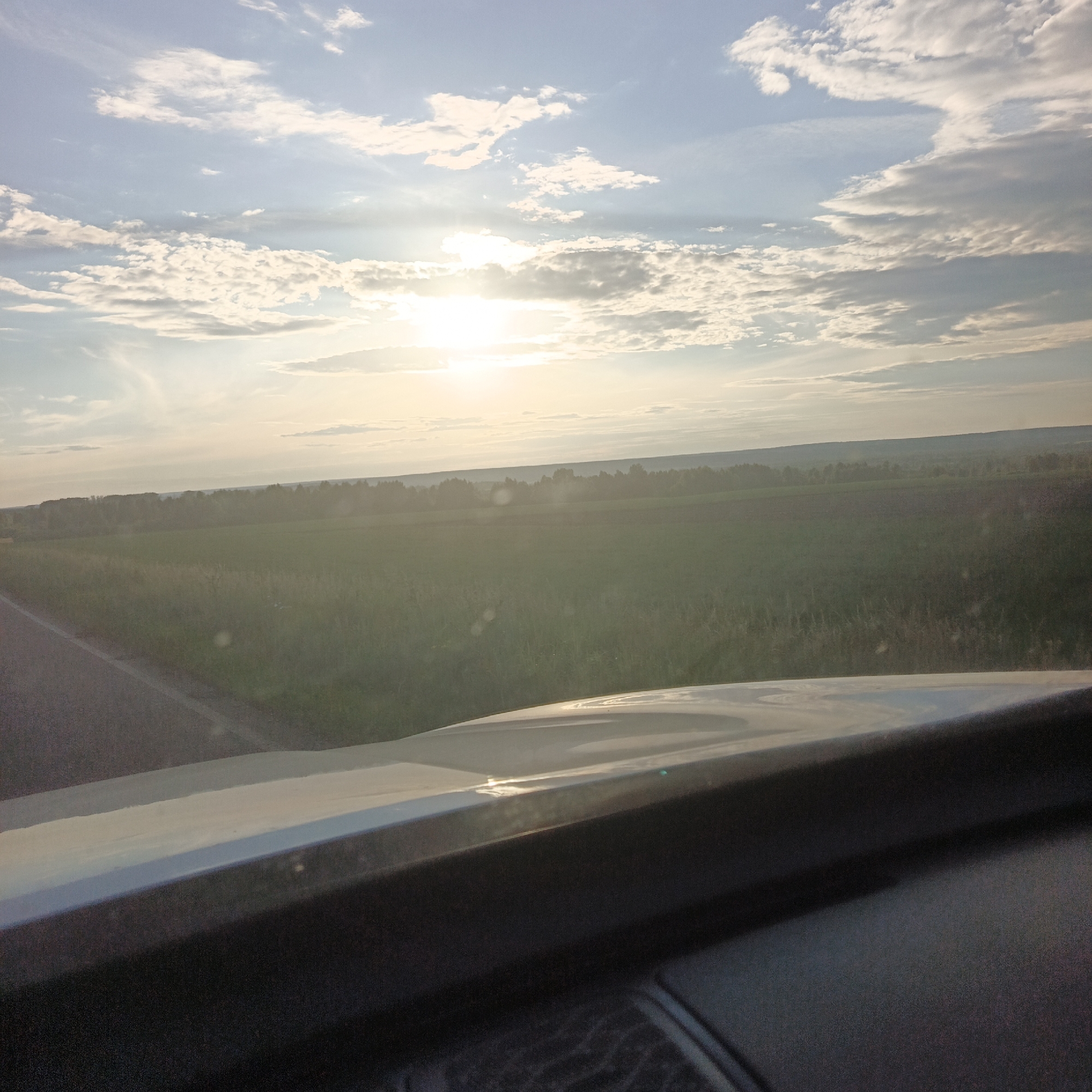 The image shows a landscape scene from the perspective of a vehicle's dashboard. In the foreground, the upper portion of a car's dashboard is visible, with a smooth, light-colored surface reflecting the sky. Beyond the dashboard, a road runs along the left side of the image, bordered by a field of tall, green grass. In the middle ground, a treeline stretches across the horizon, with a large field of what appears to be cultivated land behind it. The background is dominated by a vast sky filled with scattered white clouds. The sun is visible in the upper center of the sky, casting a bright light and creating a hazy effect over the landscape. The overall scene suggests an outdoor setting, possibly in a rural area, during daylight hours.