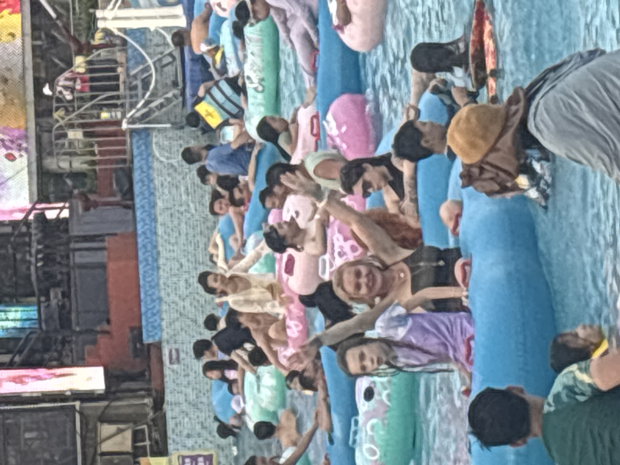 The image depicts a busy scene at an outdoor water park in Panyu District, China. Numerous individuals are seen enjoying various attractions, predominantly floating on inflatable tubes within pools of water. The environment appears to be a recreational facility with surrounding structures and decorative elements.
In the foreground, a young girl wearing a purple and white top is seated on a blue inflatable ring, looking towards the left with a surprised expression. Next to her, an adult woman with blonde hair is smiling and waving, wearing a dark tank top and a necklace. She is also on a blue inflatable. Further to the right, a person wearing sunglasses and a black top is visible, seemingly relaxed on an inflatable. In the bottom left corner, a person in a green and black t-shirt is facing away from the camera.
The middle ground shows a multitude of people of various ages, many of whom are on or around inflatable tubes in different colors, including pink and green. Interactions appear casual and relaxed, with people chatting, looking around, and enjoying the water. Some individuals are partially submerged in the water, while others are sitting on the edges of the inflatables.
In the background, there are more pools and structures characteristic of a water park, including what appears to be a slide or elevated platform with railings. A person in a yellow and blue life vest is visible, suggesting ongoing activities and supervision. The overall atmosphere is one of leisure and fun. The water is a prominent element, reflecting light and creating a vibrant blue hue. The lighting suggests it is daytime.