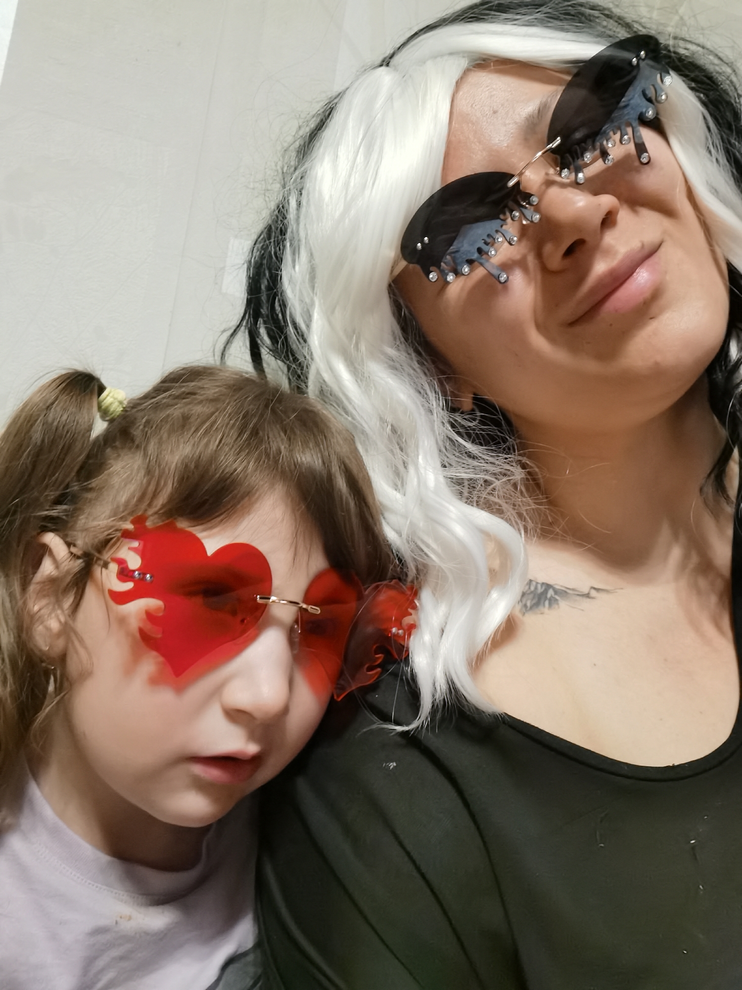 A medium shot depicts a woman and a child posing for a selfie. The child, with light brown hair in pigtails and wearing red heart-shaped sunglasses with flame details, leans against the woman. The woman, dressed in black, has a black and white wig and oversized black sunglasses with crystal embellishments. A tattoo is visible on her upper chest. The background appears to be a plain wall. There is no clear indication of being in Votkinsk, Russia.