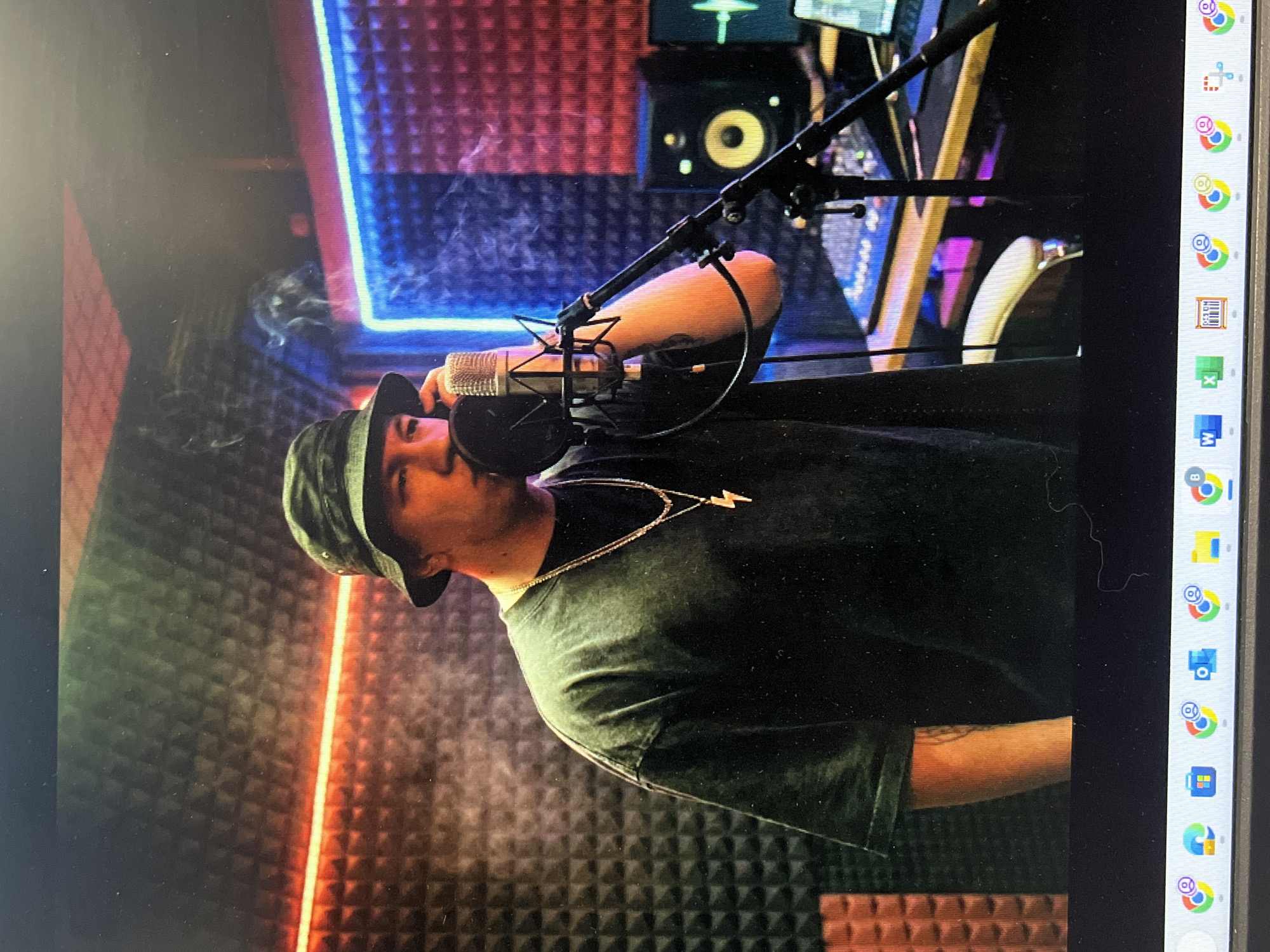 In a recording studio, a man wearing a bucket hat and a dark t-shirt stands before a microphone, appearing to be singing or rapping. Smoke billows around him, illuminated by the red and blue neon lights lining the sound-dampened walls. The man wears two layered silver necklaces, one with a lightning bolt pendant. In the background, audio equipment, including speakers and a mixing board, is visible, along with a computer screen displaying audio waveforms. The image is a photograph taken from a screen, as indicated by the visible computer taskbar at the bottom. There is no information in the image to suggest the location is in Lupanovo, Russia.
