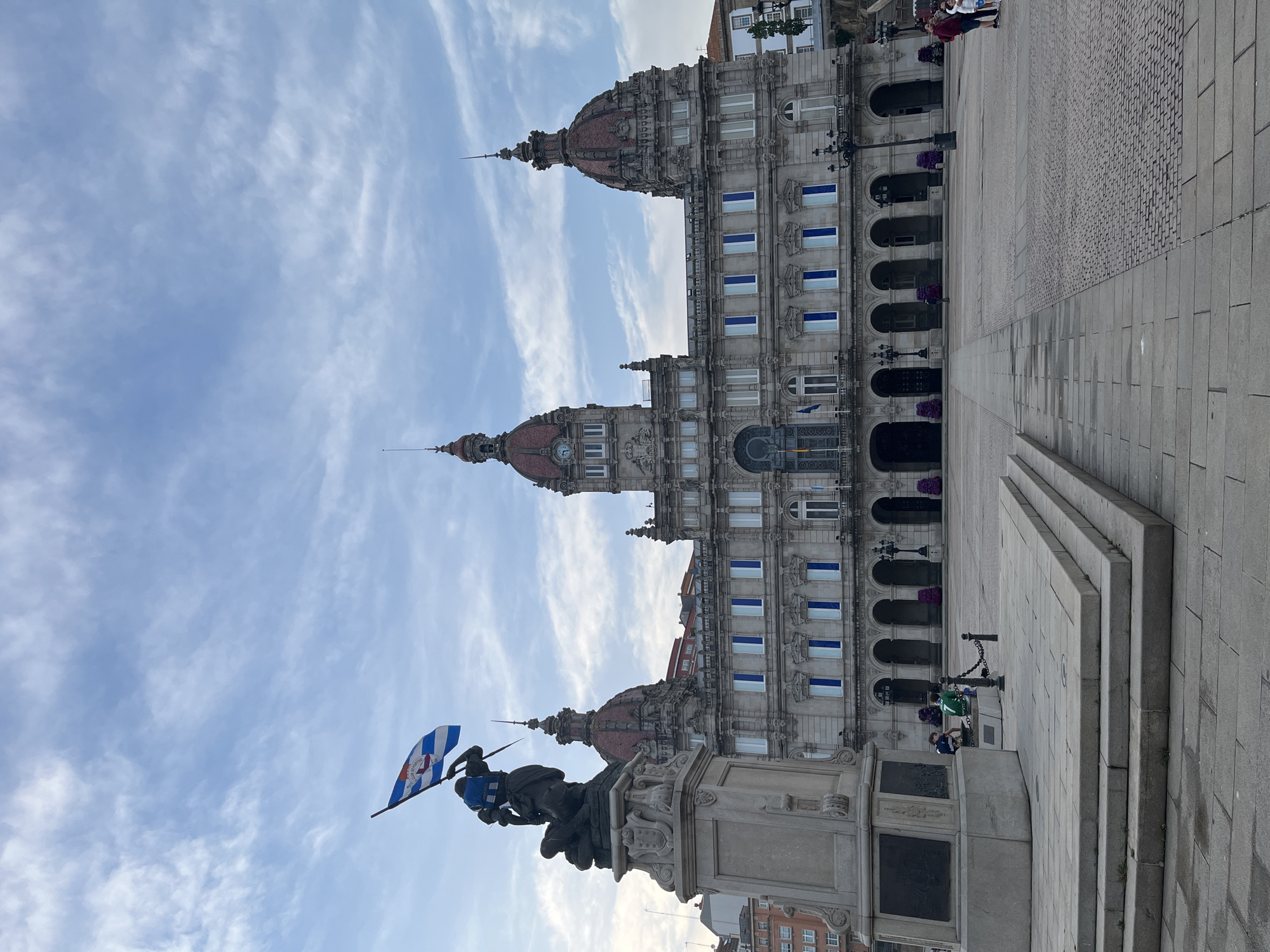 The image captures a view of the María Pita Square in A Coruña, Spain, featuring the María Pita statue in the foreground and the A Coruña City Hall in the background. The statue is a monument of María Pita, a local heroine. It consists of a bronze figure holding a flag. The flag features a blue and white diagonal pattern with a crest. The statue rests on a stone pedestal, which has inscriptions on its sides.
The City Hall is a large, ornate building with three domed towers. It features several rows of arched windows and balconies, adorned with decorative elements. The building's facade is light-colored, and the domes appear to be made of red tiles.
The sky above is a mix of blue and white, with wispy clouds scattered throughout. The ground is paved with stone slabs, and there are a few people visible in the background. Some people are walking nearby, and a few are sitting on the steps in front of the statue.