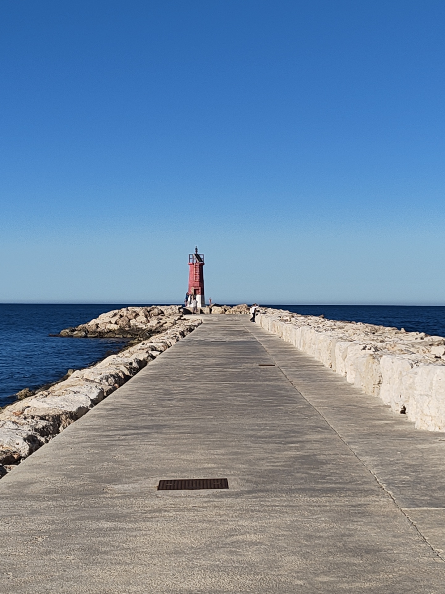 A long concrete pier extends straight into the deep blue sea under a perfectly clear, cerulean sky in Dénia, Spain. The pier, made of light grey concrete, is bordered on both sides by robust barriers of large, light-colored, rough-hewn rocks. Embedded in the concrete surface are several rectangular metal grates, with one clearly visible in the foreground.
At the far end of the pier, a prominent red lighthouse with a white top and a visible ladder or staircase stands tall. Two individuals are present near the lighthouse; one stands closer to its base, while another is partially obscured further back. Additionally, another person wearing a white top and dark trousers is seen walking away from the viewer along the right side of the pier, roughly midway between the foreground and the lighthouse.
The sea appears calm with gentle ripples, reflecting the bright sky. The overall weather is sunny and clear, indicative of a pleasant day, likely late morning or early afternoon given the bright illumination. The scene conveys a sense of tranquility and offers a peaceful view of the coastal environment. There is no visible text in the image.