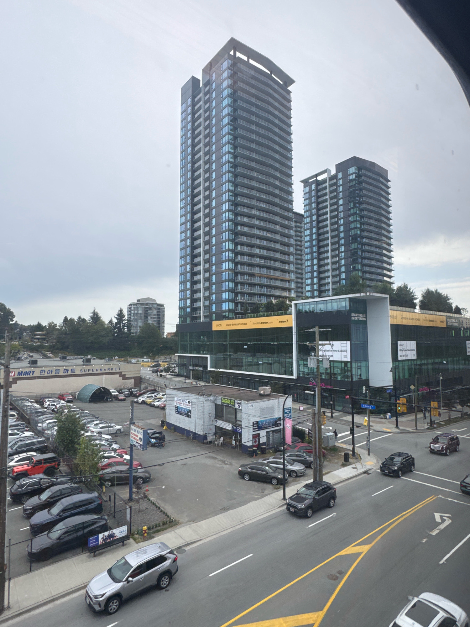 The image depicts a street view in Burnaby, Canada, with a focus on urban development and vehicle traffic. Dominating the skyline are two modern, tall residential buildings with glass facades, likely condominiums, with a smaller, older grey building visible behind them. In the foreground, a busy street with multiple lanes is observed, with various cars driving or parked. Yellow lines demarcate the lanes and turn-only paths. A variety of vehicles are visible, including sedans, SUVs, and a red jeep.
Several commercial establishments are present. A low-rise building with signage in English and Korean, "MART and H MART," indicates a supermarket. Adjacent to this, a large parking lot is filled with numerous vehicles. Further down the street, another commercial building features large glass windows and prominent signage for "SOCO" and "MOVE-IN READY HOMES" with an "Anthem" logo, suggesting a real estate development or sales office. Other businesses like "LUBE KING" and "ZAGROS" are also visible on signage for smaller establishments.
The environment appears to be overcast, with a cloudy sky. Trees are scattered throughout the scene, particularly behind the low-rise commercial buildings and between the residential towers, suggesting a suburban or mixed-use urban area. The street is well-maintained, with sidewalks, traffic lights, and street signs, including one indicating "Young." There are a few pedestrians visible on the sidewalks, suggesting a relatively active urban environment.